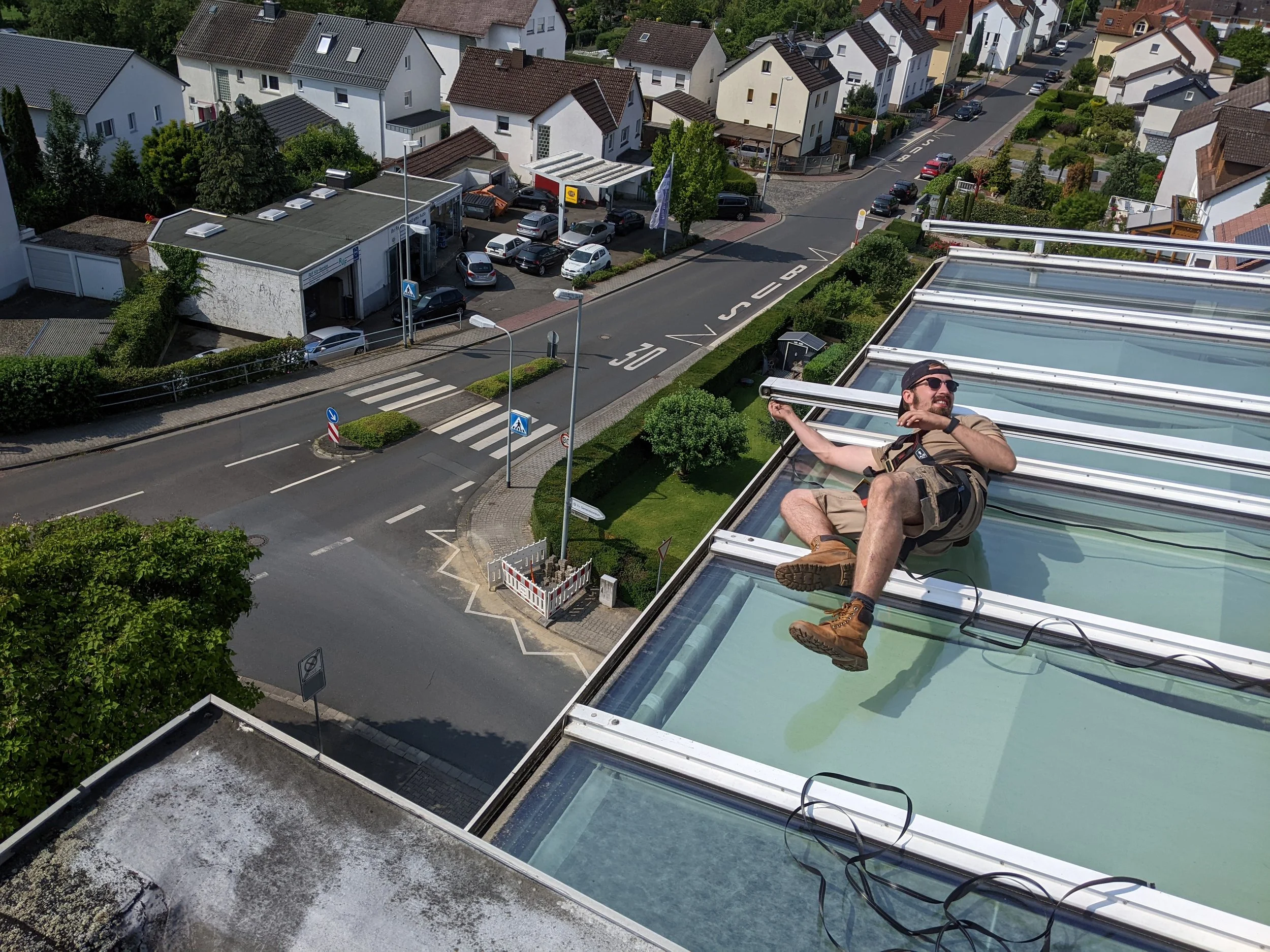 Mann liegt auf dem Dach eines Gebäudes auf einer Solar- oder Photovoltaikanlage, während er an den Sonnenkollektoren arbeitet. Er trägt Arbeitskleidung, Sonnenbrille und sitzt auf den transparenten Abdeckungen der Anlage, mit einer Stadtansicht im Hi