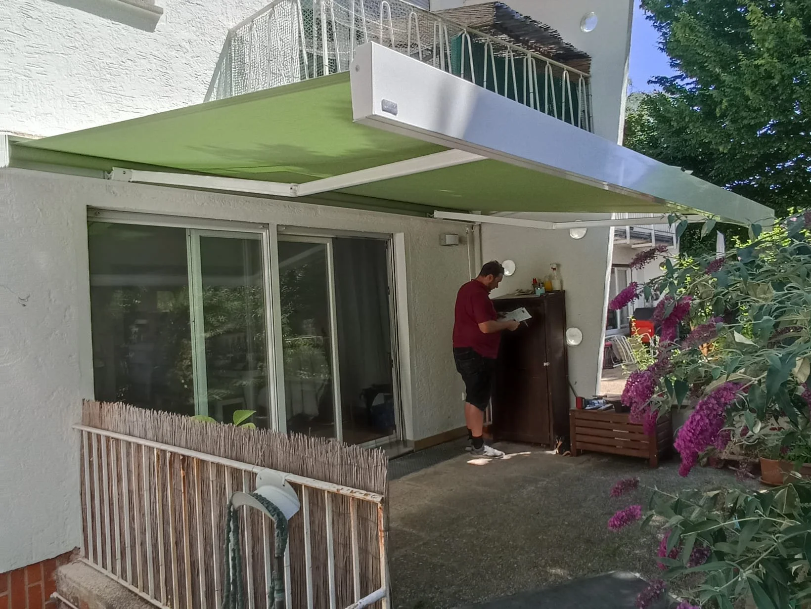 Ein Mann steht vor einer weißen Hauswand und liest in einer Zeitschrift oder einem Katalog. Es gibt einen kleinen Garten mit bunten Blumen auf der rechten Seite, eine Glas-Schiebetür und einen kleinen Balkon oben.