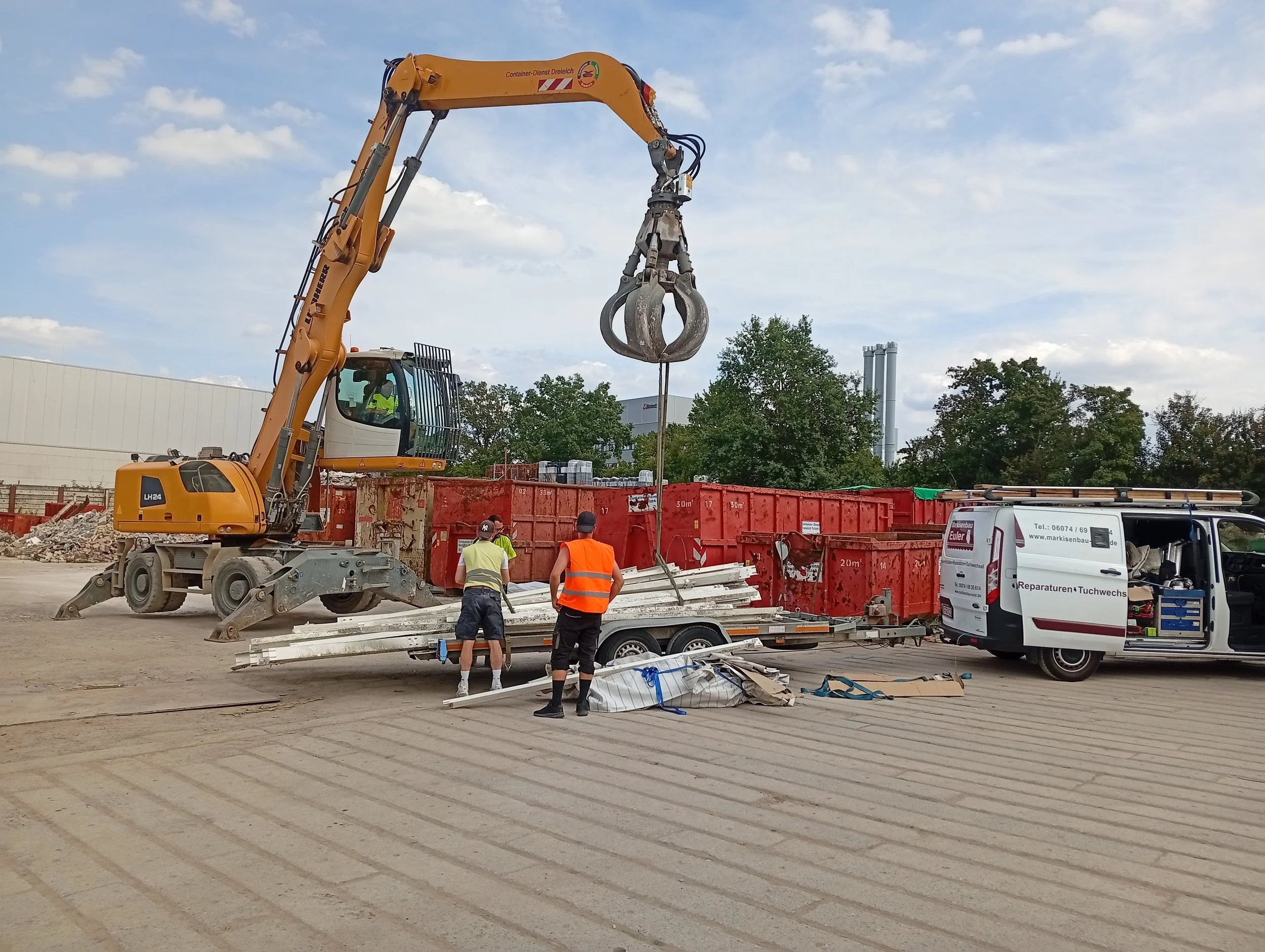 Arbeitscrew beim Baustellenaufbau mit Kran, der Baumaterialien hebt, auf einer Baustelle mit Bauwagen, Baugerüsten und Bäumen im Hintergrund, bei Tageslicht.
