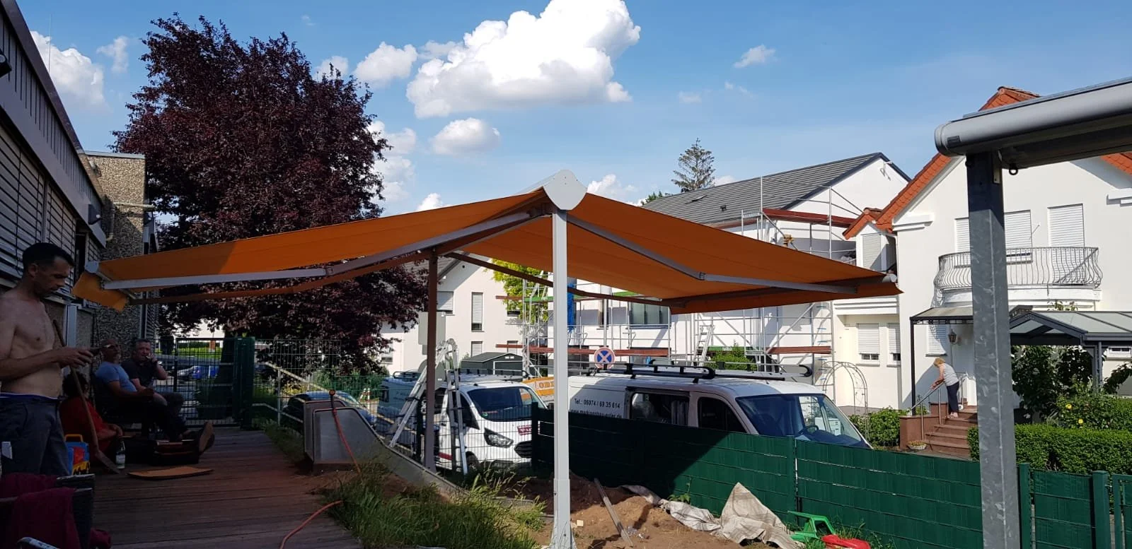 Ein Baustellenszene mit einem orangefarbenen Sonnensegel, mehreren Personen, einem weißen Lieferwagen und einem Haus im Hintergrund bei schönem Wetter.
