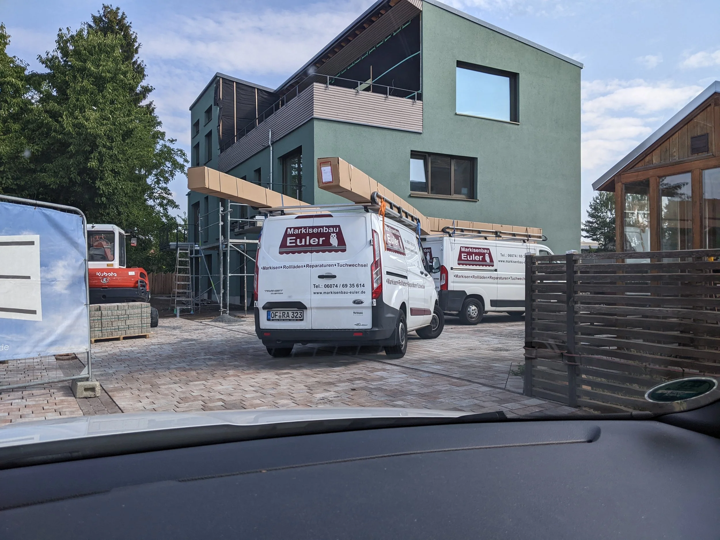 Baustelle mit zwei weißen Firmenautos, einem grünen Haus im Hintergrund, Baugerüsten und Baustoffen.