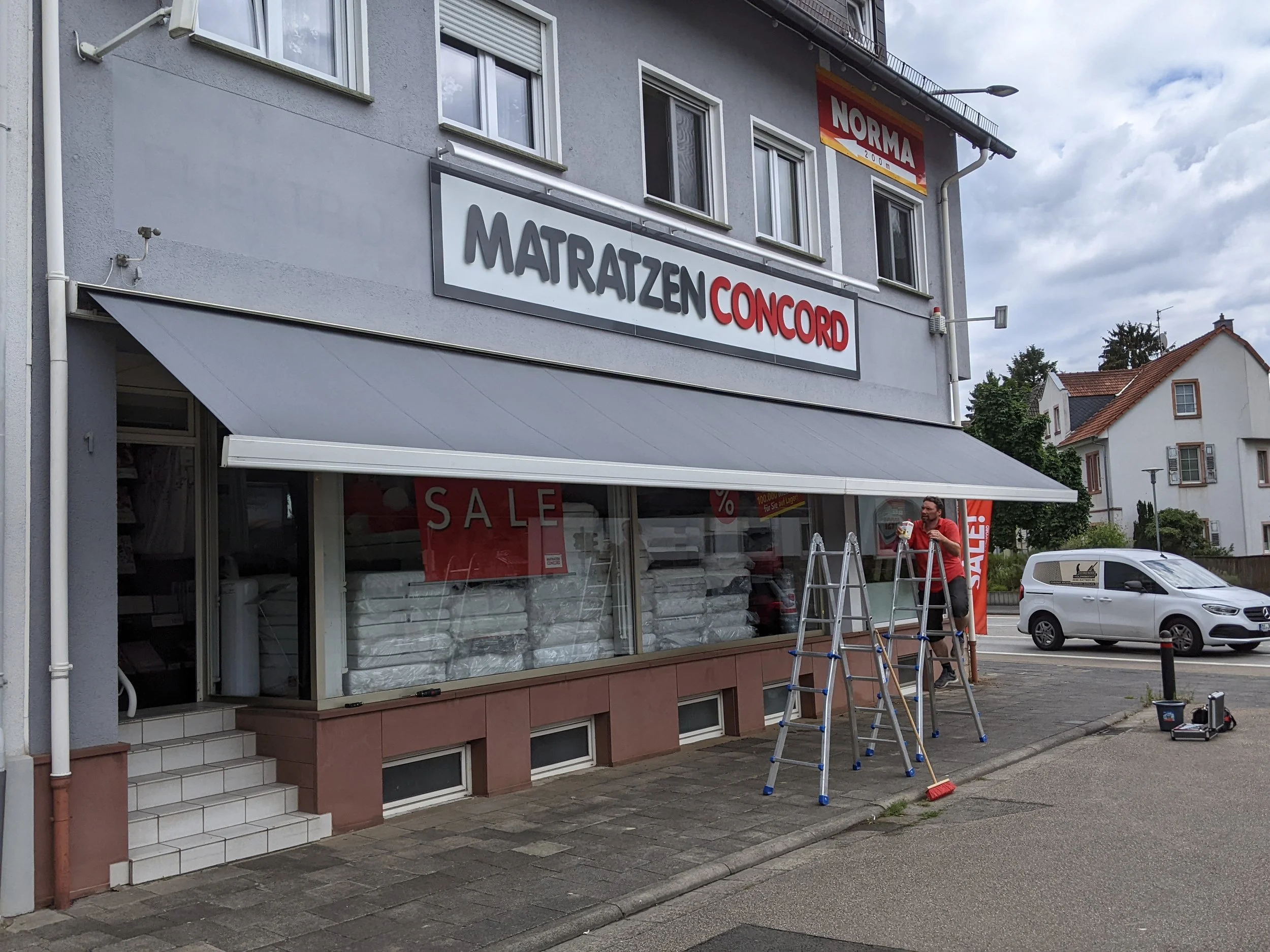 Ein Mann steht auf einer Leiter vor einem Geschäft, das Matratzen verkauft. Es gibt einen großen Verkaufsaufsteller mit dem Wort 'SALE' im Fenster.