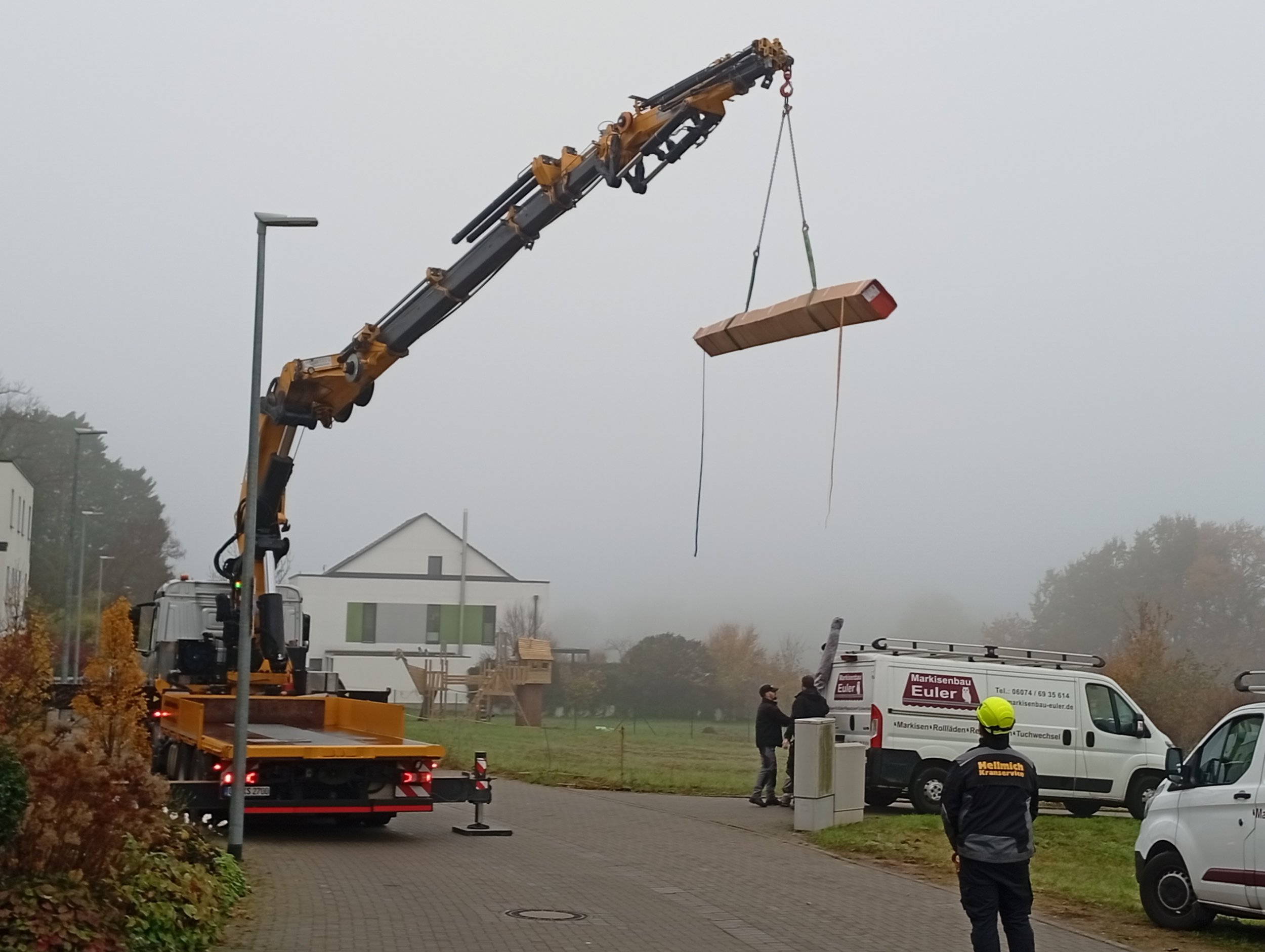Ein Kran hebt ein Holzstück in einer Wohngegend bei Nebel, mit mehreren Personen und Firmenfahrzeugen im Einsatz.
