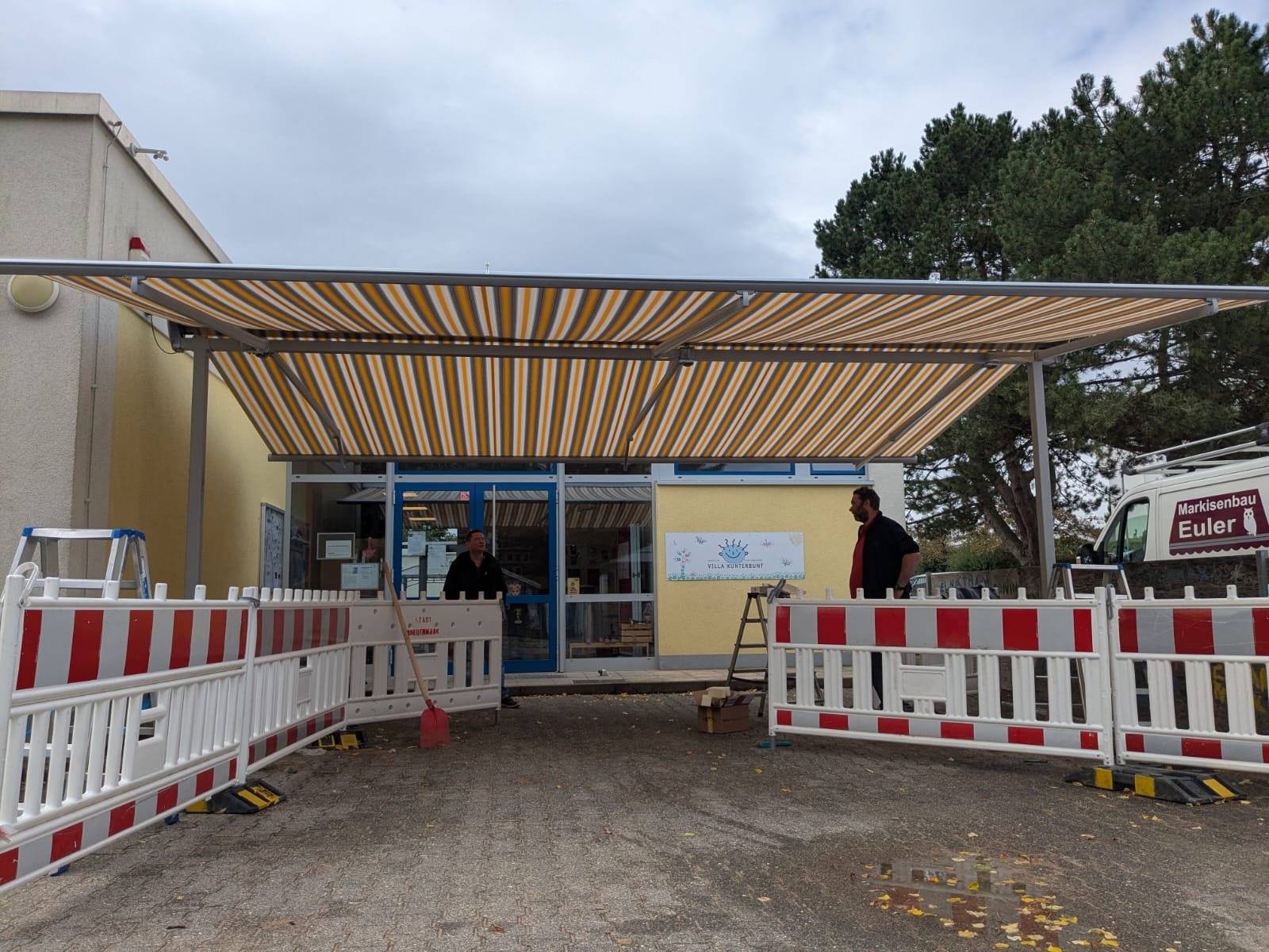 Baustelle vor einem Gebäude mit gelben Wänden und bedrucktem Sonnenschutz, Bauarbeiter arbeiten, Absperrungen chevroniert, ackerige Oberfläche, hinter Bäumen.