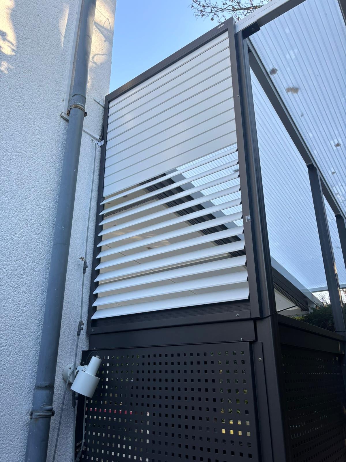 Außenansicht einer modernen Terrasse mit weißen verstellbaren Lamellen und einer Glaswand, teilweise sichtbar an einer weißen Hauswand.