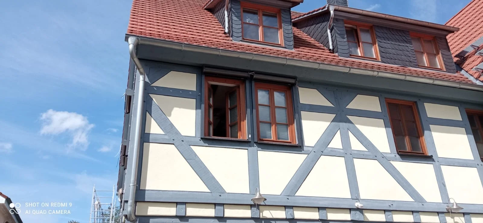Ein Haus im Fachwerkstil mit weißen Wänden, blauen Holzträgern und roten Doppelfenstern, Dach mit roten Ziegeln und mehreren Fenstern.