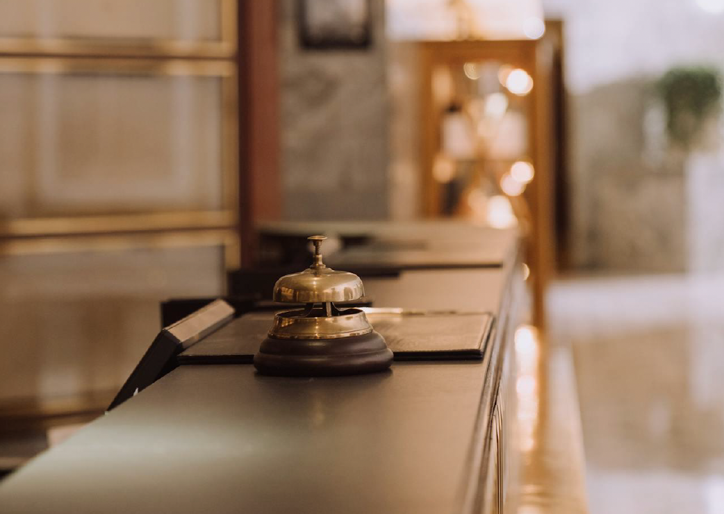 A hotel reception desk with a bell and a folder, in a warmly lit lobby with wooden and stone interior design.