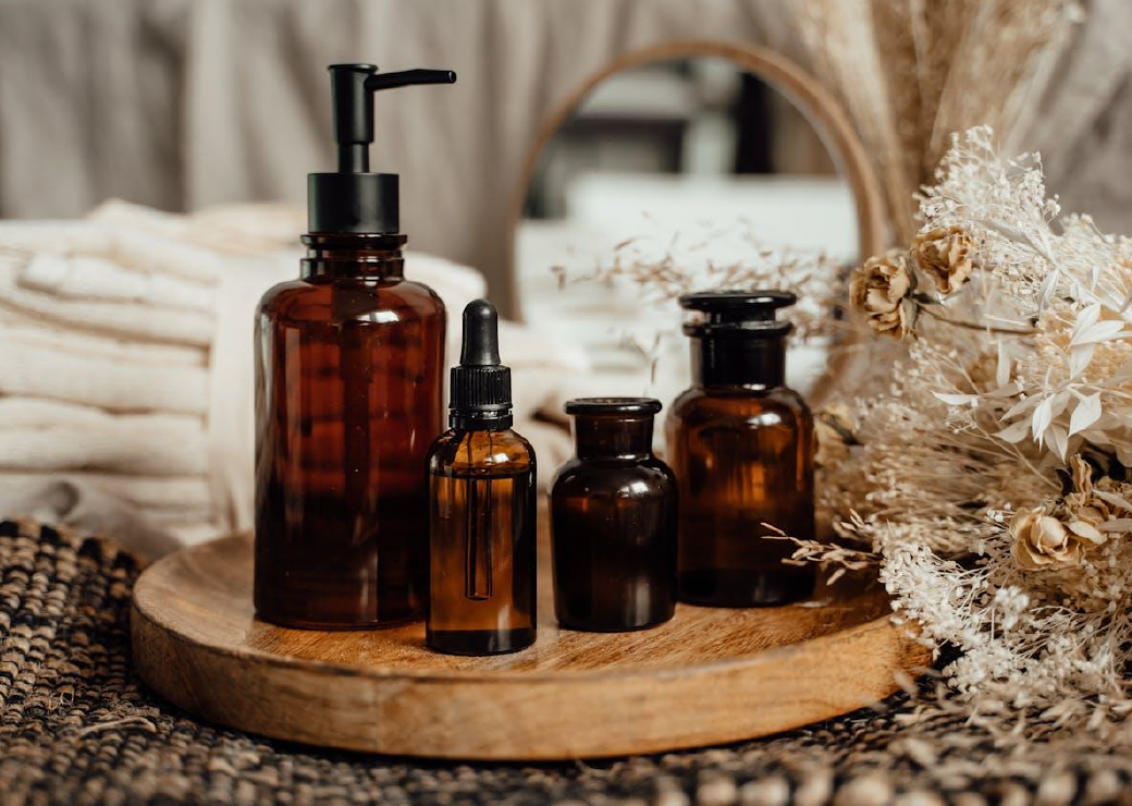 Four amber glass bottles of different sizes on a wooden tray, with dried flowers and a mirror in the background.
