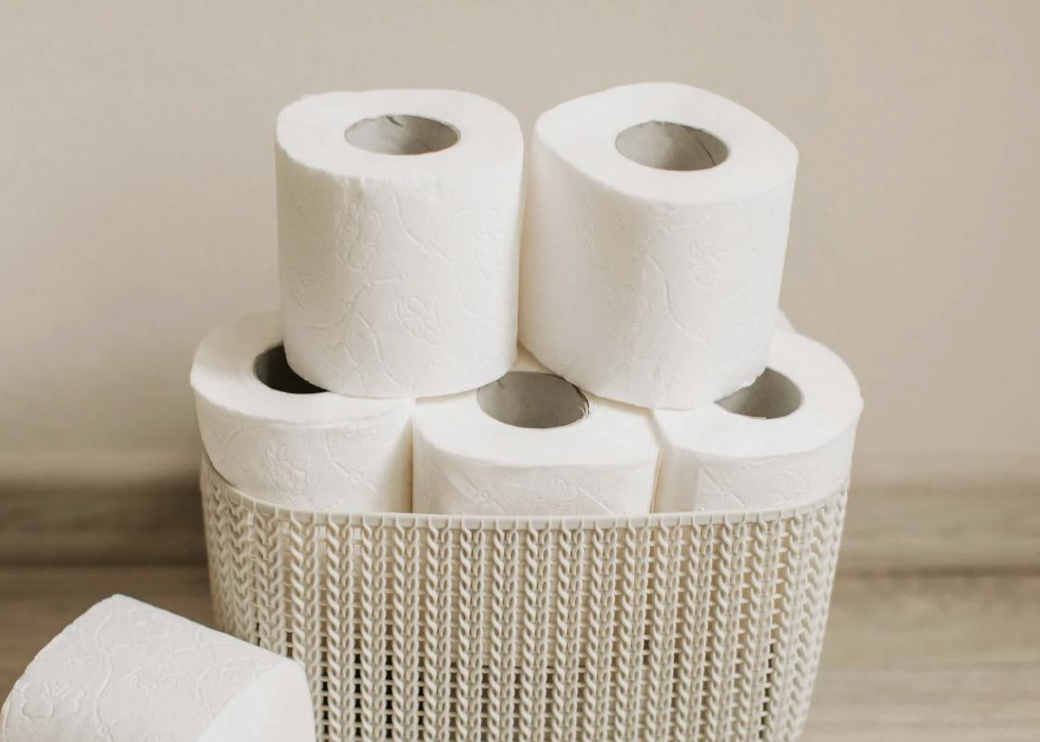 Six white toilet paper rolls arranged in a wooden holder with a textured design, against a neutral wall and wooden surface.