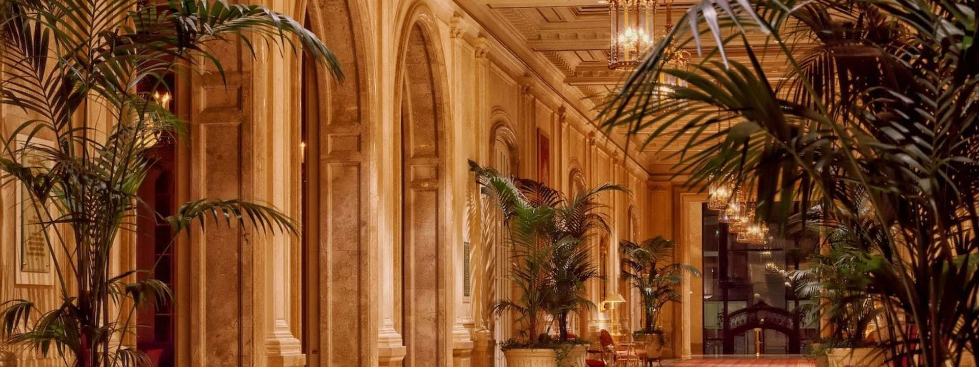 Luxurious hotel lobby with marble walls, tall arched windows, lush green plants in large pots, chandeliers, and elegant furnishings.