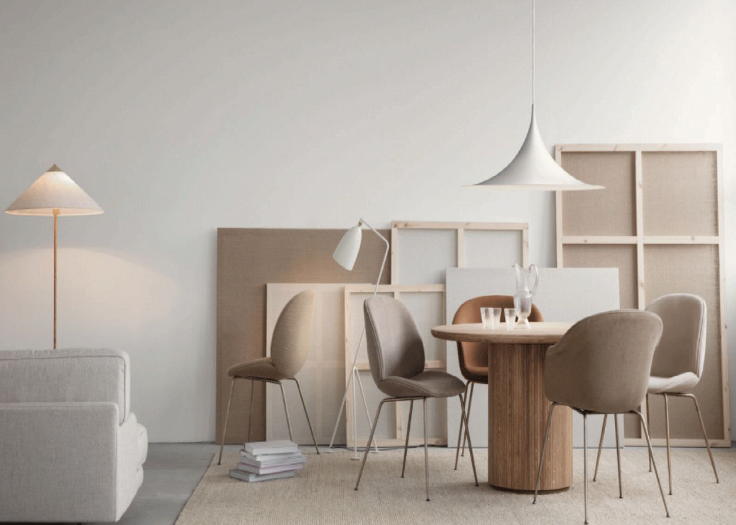 A minimalist living room with a round wooden table, upholstered chairs, a white sofa, a standing and pendant light, and several picture frames leaning against the wall.