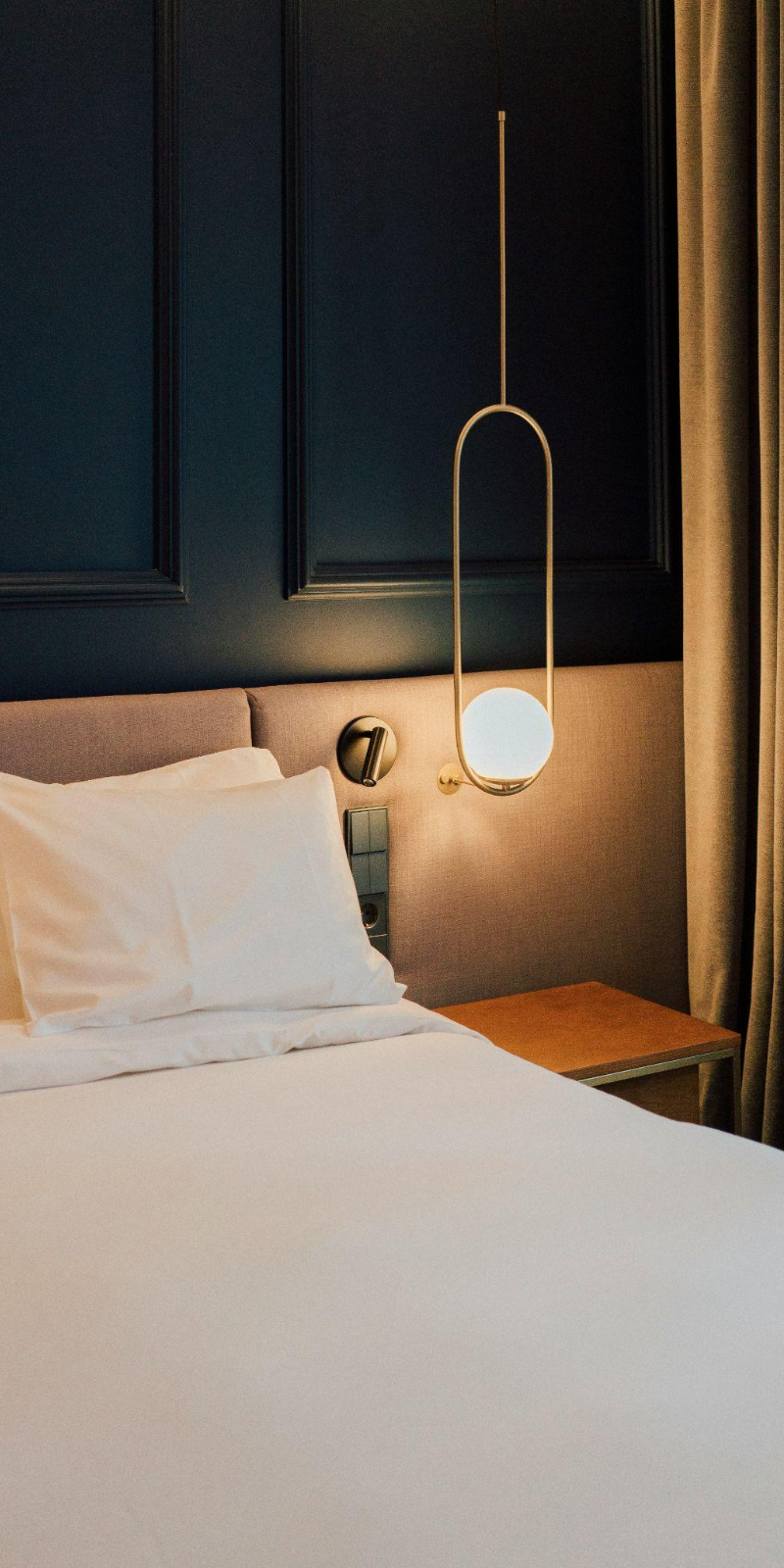 Close-up of a hotel bed with white sheets and pillows, a beige headboard, and an elegant hanging bedside lamp with a round white globe. The wall behind the bed features dark blue paneling, and a small wooden bedside table is visible to the right.