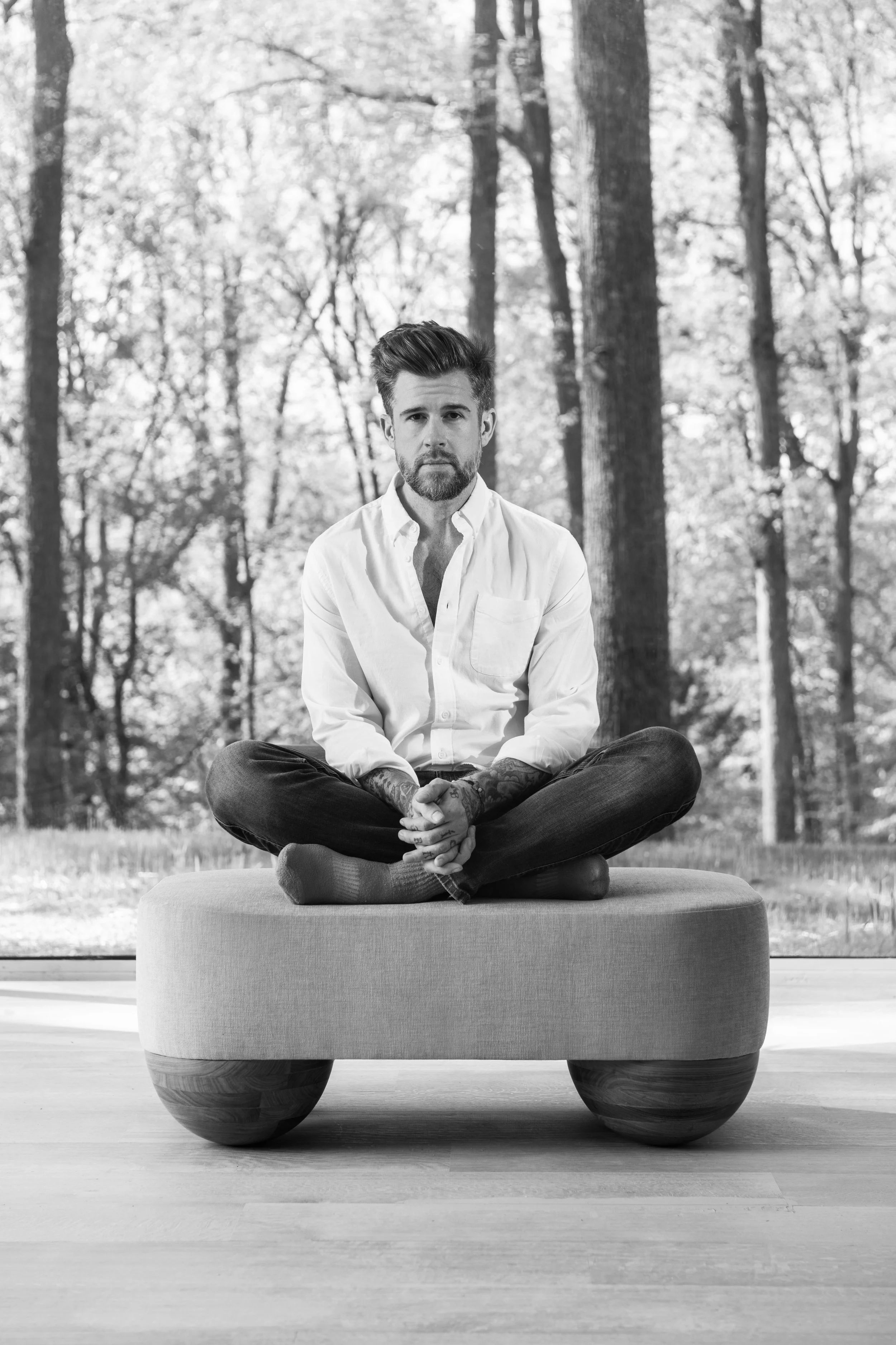 A man with a beard and tattoos, wearing a white shirt and dark jeans, sitting cross-legged on a padded platform in a room with a large window showing trees outside.