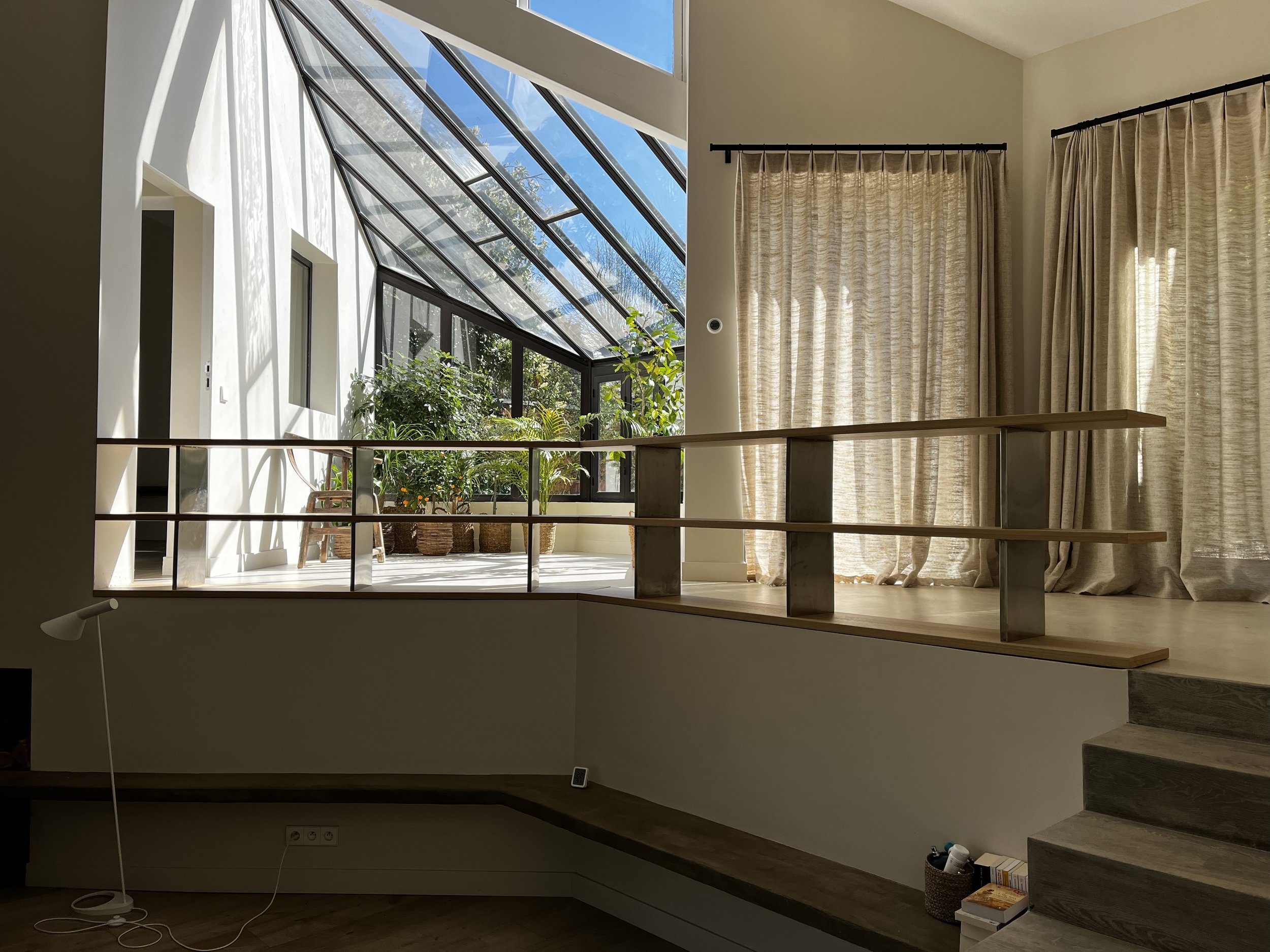 Intérieur d'une maison avec un grand panneau de fenêtre en verre inclinée permettant la lumière naturelle, des plantes dans le coin, et des rideaux clairs.