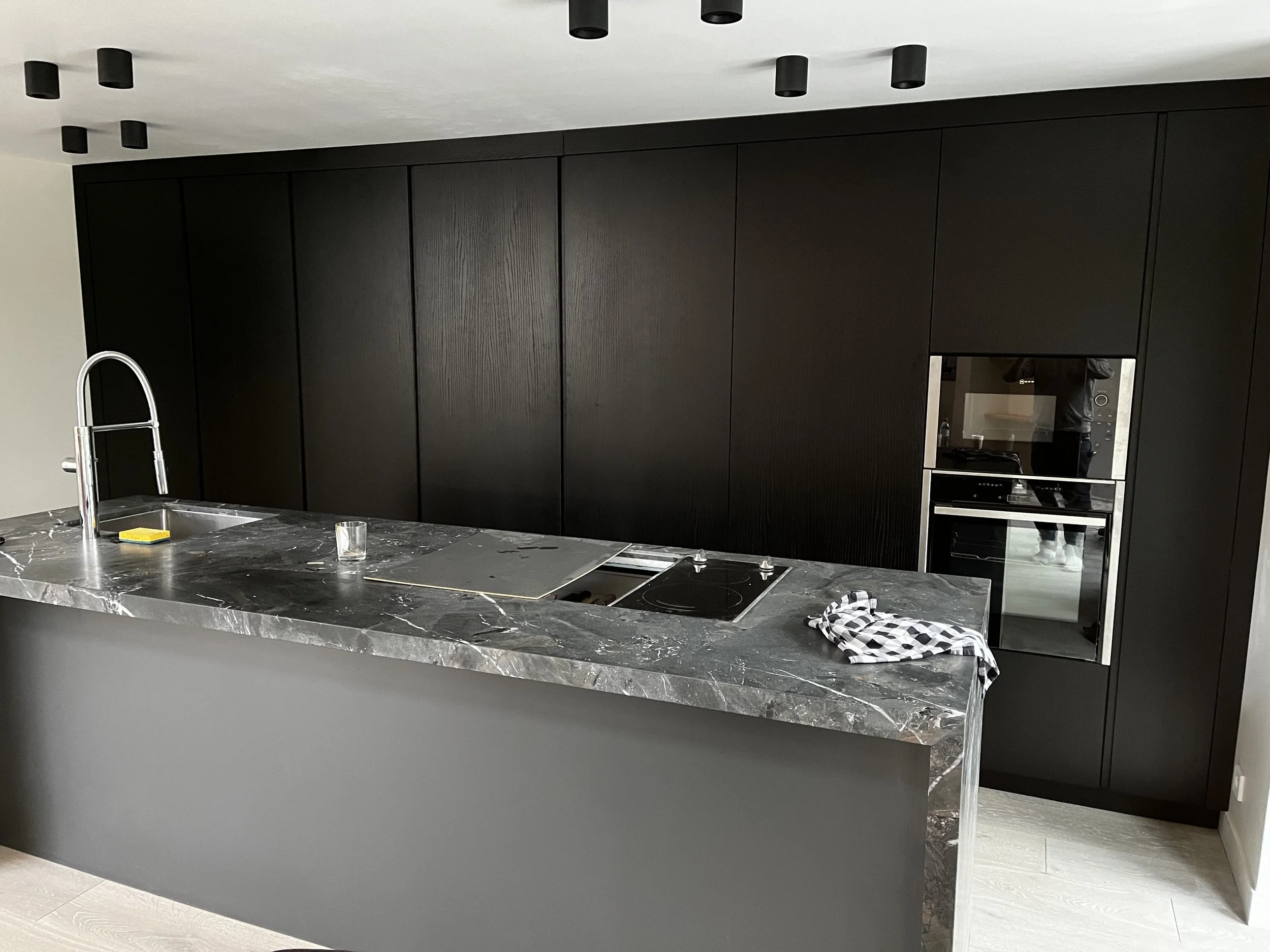 Cuisine moderne avec un grand plan de travail en marbre noir et un placard noir du sol au plafond. Une évier en acier inoxydable avec robinet est visible sur le plan de travail. Il y a une plaque de cuisson électrique ou à induction et une porte inté