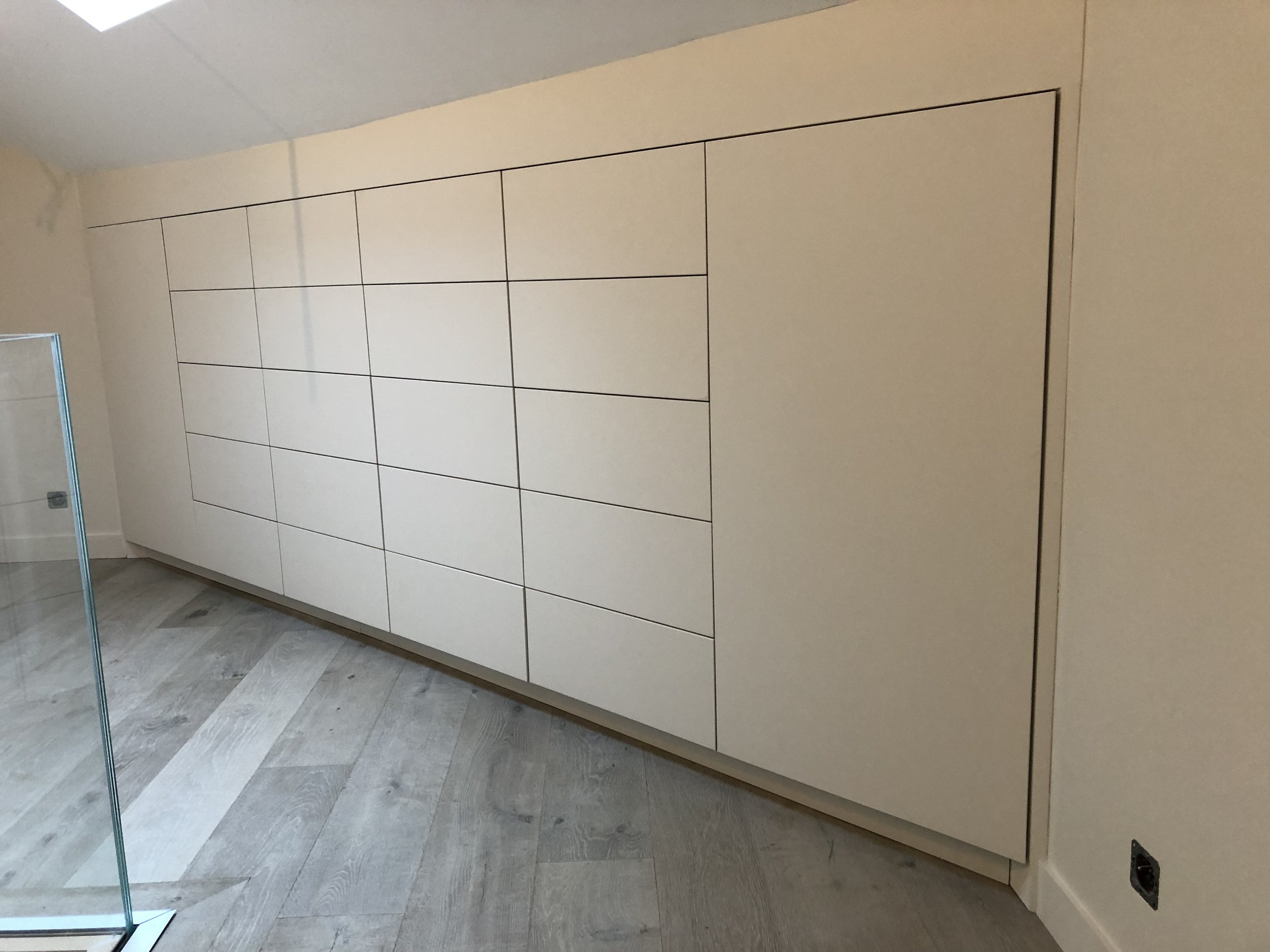 Grand placard blanc avec de nombreuses portes dans une pièce blanche, sol en carrelage gris, mur beige, avec prise électrique visible en bas à droite.