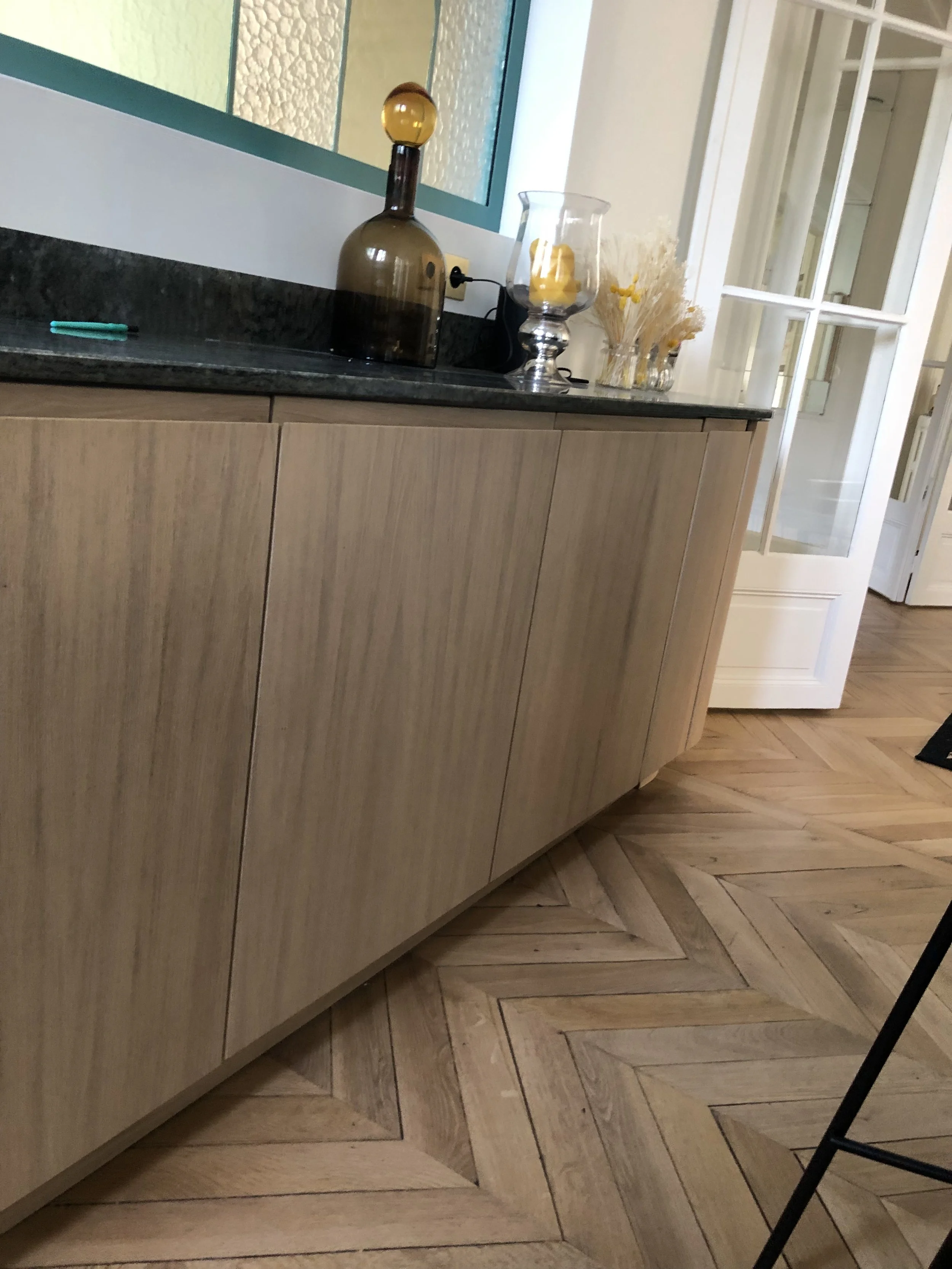 Un meuble en bois clair avec un plan en marbre noir, décoré avec une grande bouteille en verre teinté marron, une lanterne en verre, et un bouquet de fleurs séchées dans un vase."