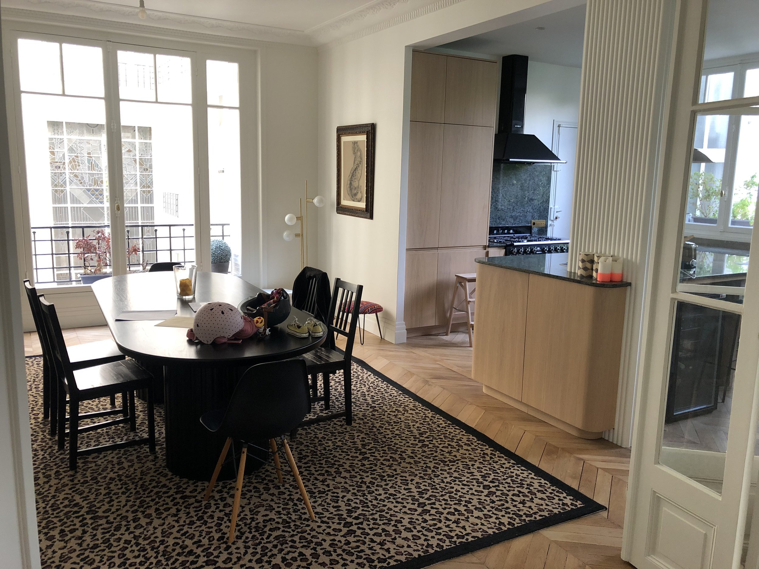 Salle à manger avec table ronde, chaises noires, tapis à motif léopard, cuisine en arrière-plan, fenêtres grandes laissant entrer la lumière, décoration intérieure moderne.