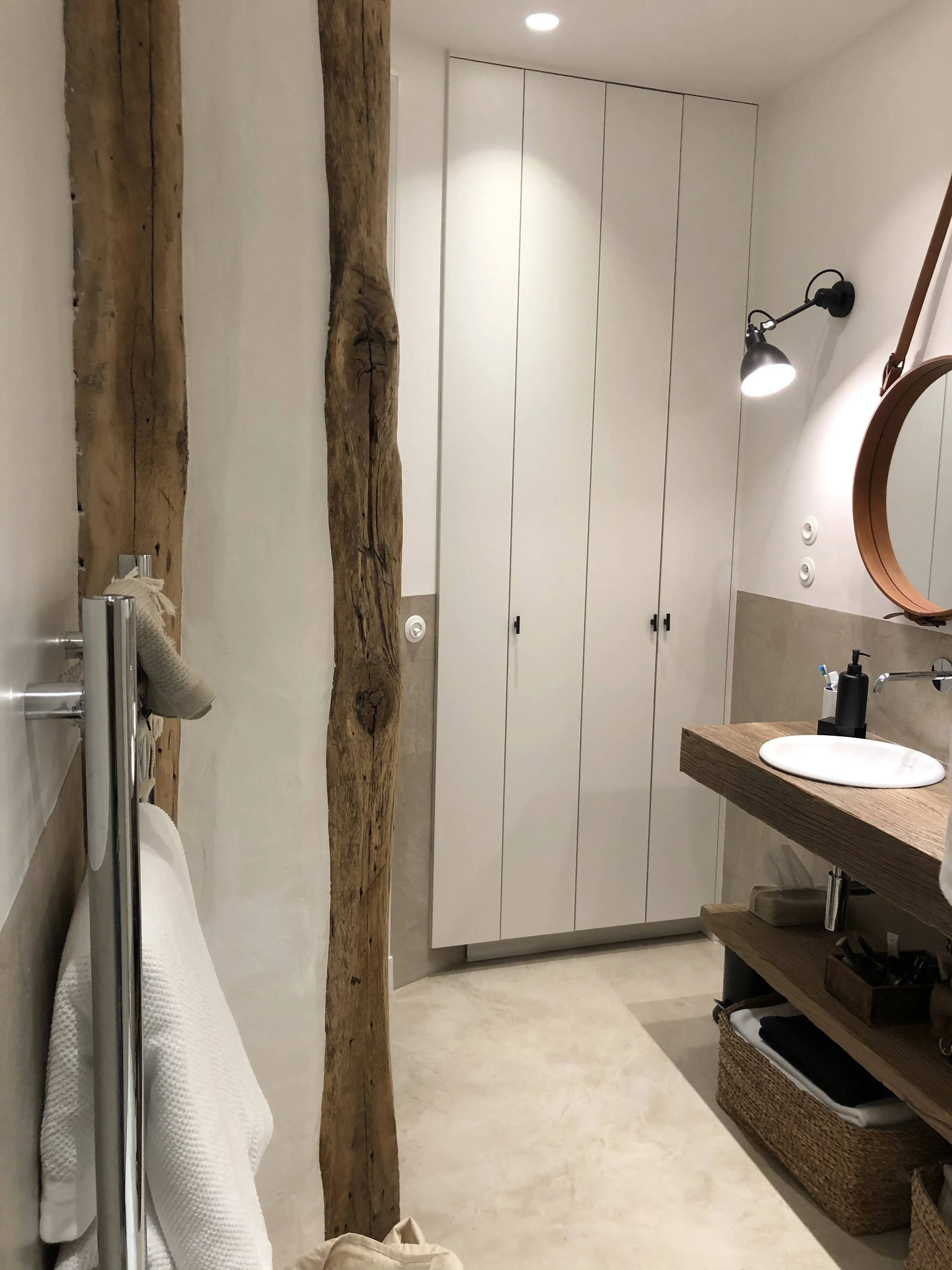 Ceinture de salle de bain en toile, serviettes blanches, poutre en bois brut, meubles de rangement en bois naturel, miroir rond, lavabo en céramique, lampes murales.