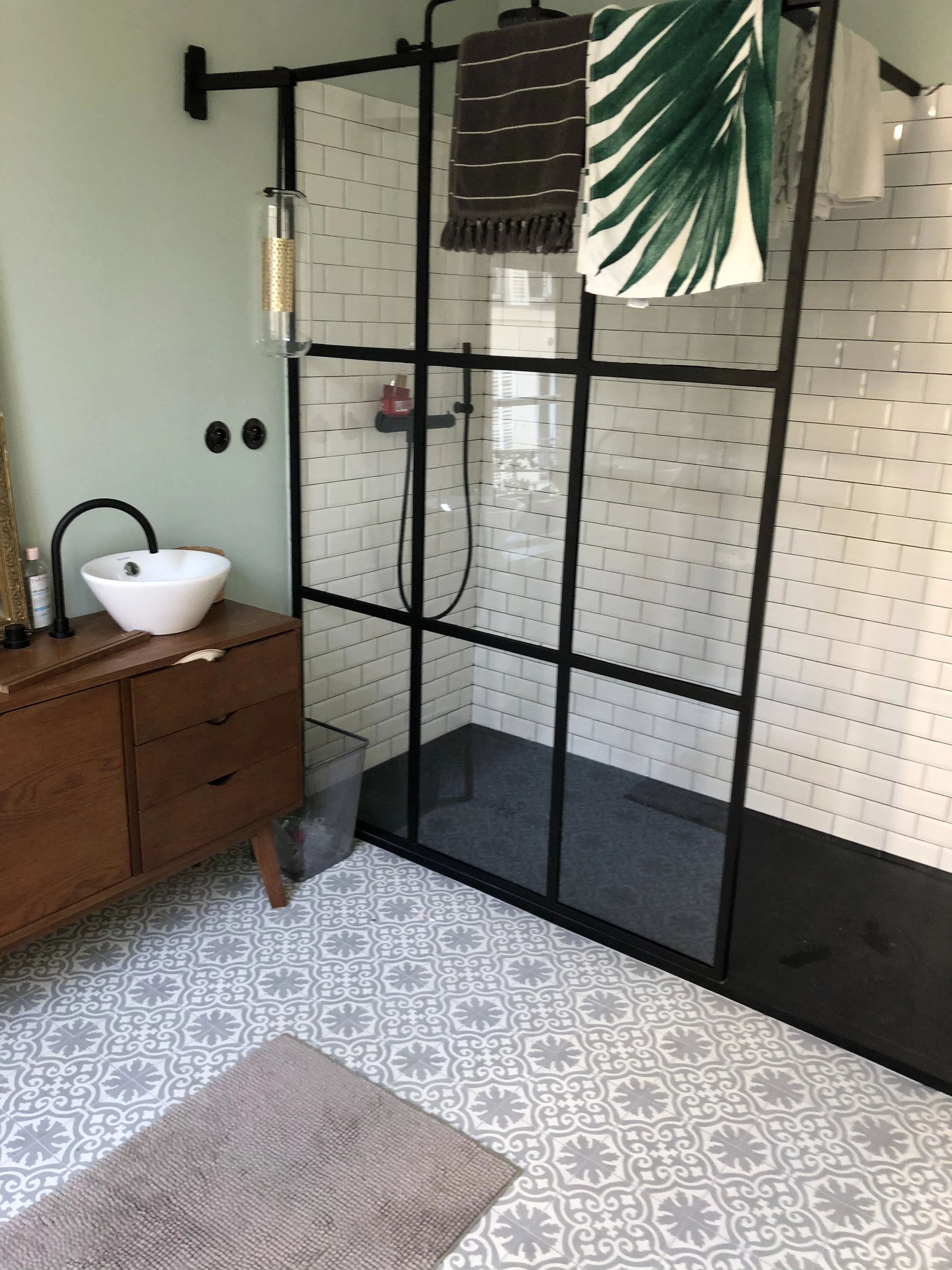 Salle de bain avec une cabine de douche noire, un lavabo rond blanc sur un meuble en bois, un miroir, deux prises électriques et un tapis beige. Le sol a un motif en carreaux gris et blancs.