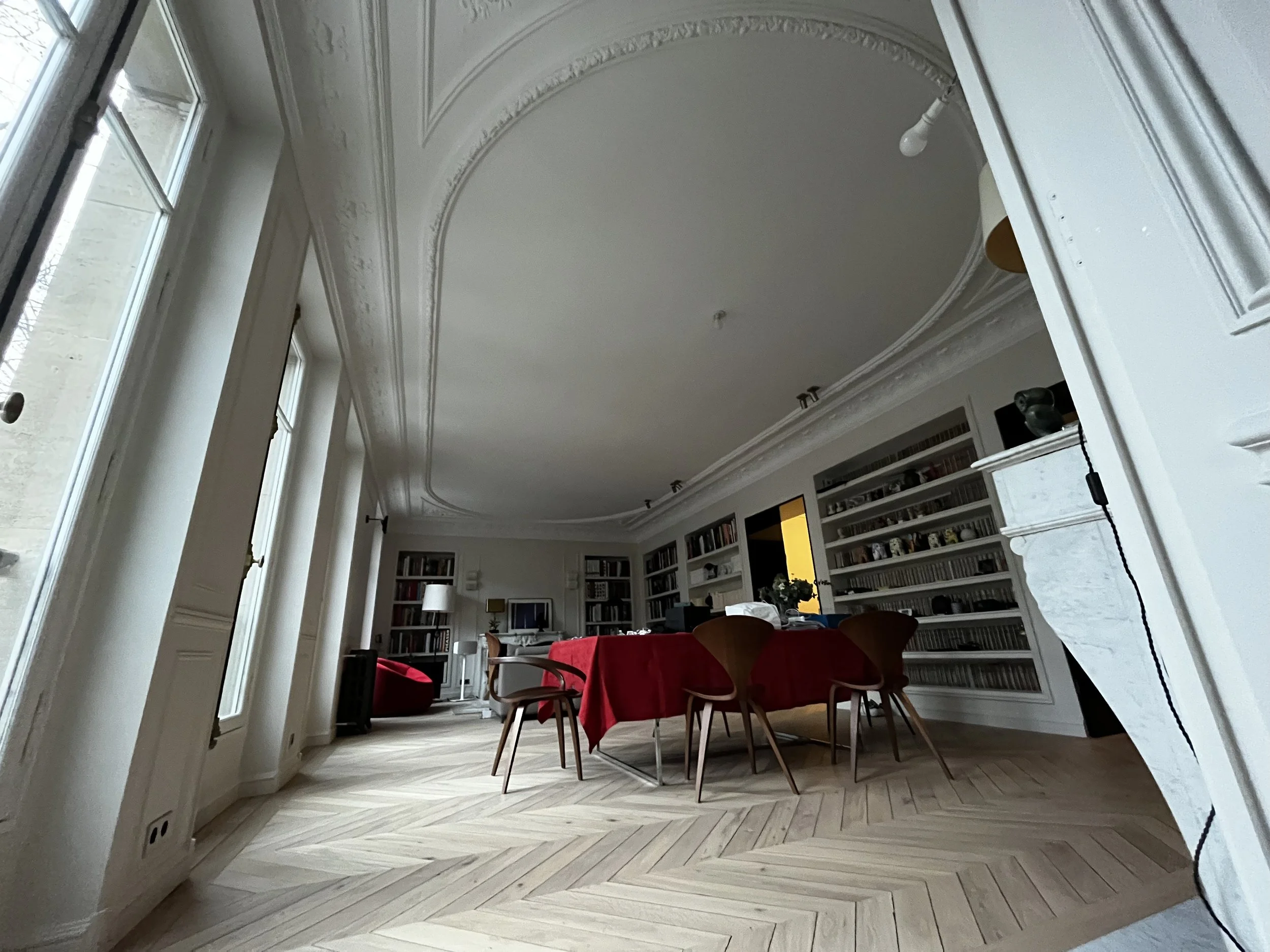 Intérieur d'une pièce élégante avec un plafond orné de moulures, grandes fenêtres laissant entrer beaucoup de lumière, une bibliothèque remplie de livres, une table avec une nappe rouge et des chaises en bois.