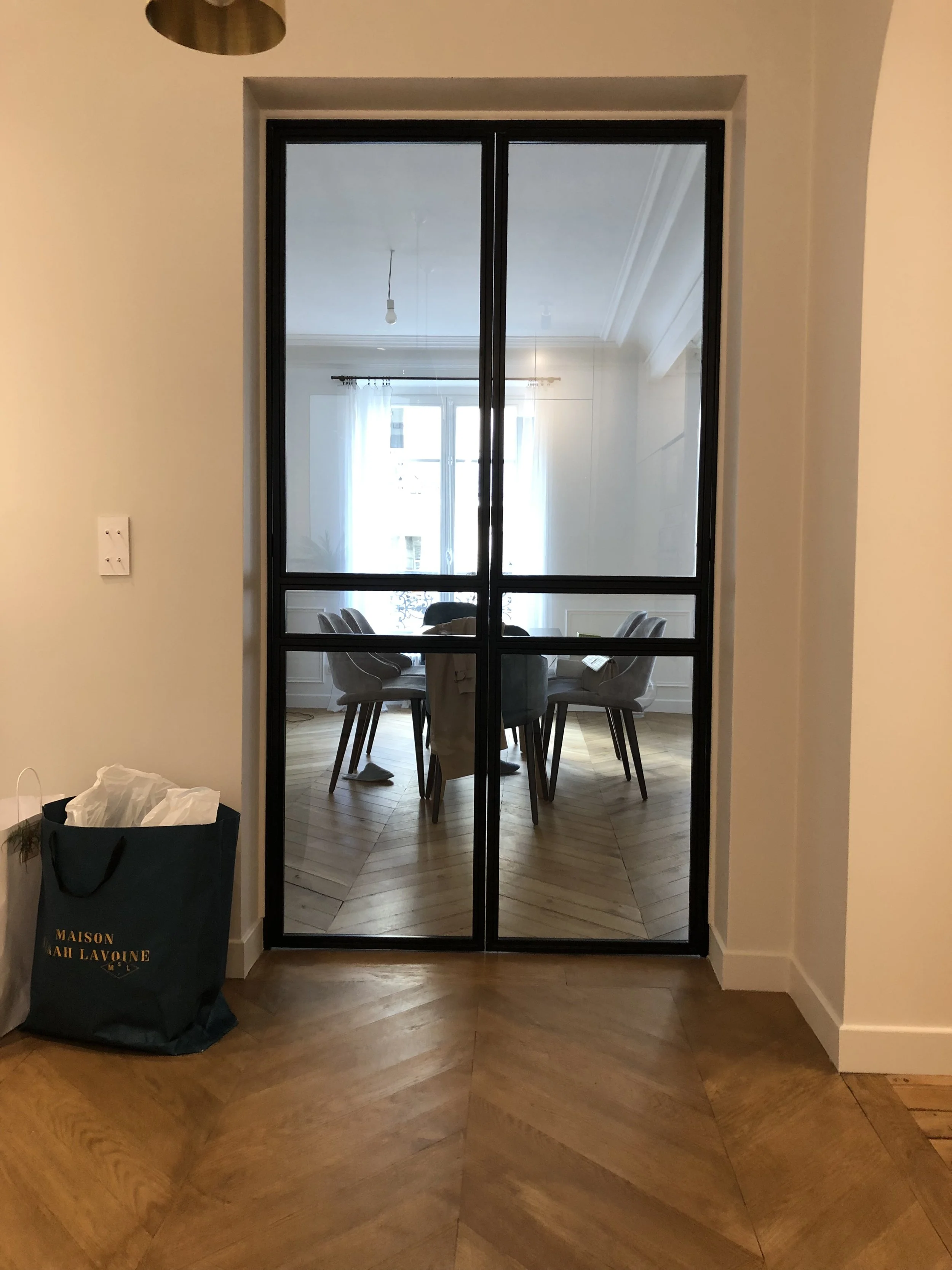 Une porte vitrée noire donnant sur une salle à manger avec une table ronde et des chaises, vue depuis un hall avec un sol en parquet en chevrons. À gauche, un sac en tissu noir avec des écritures dorées.