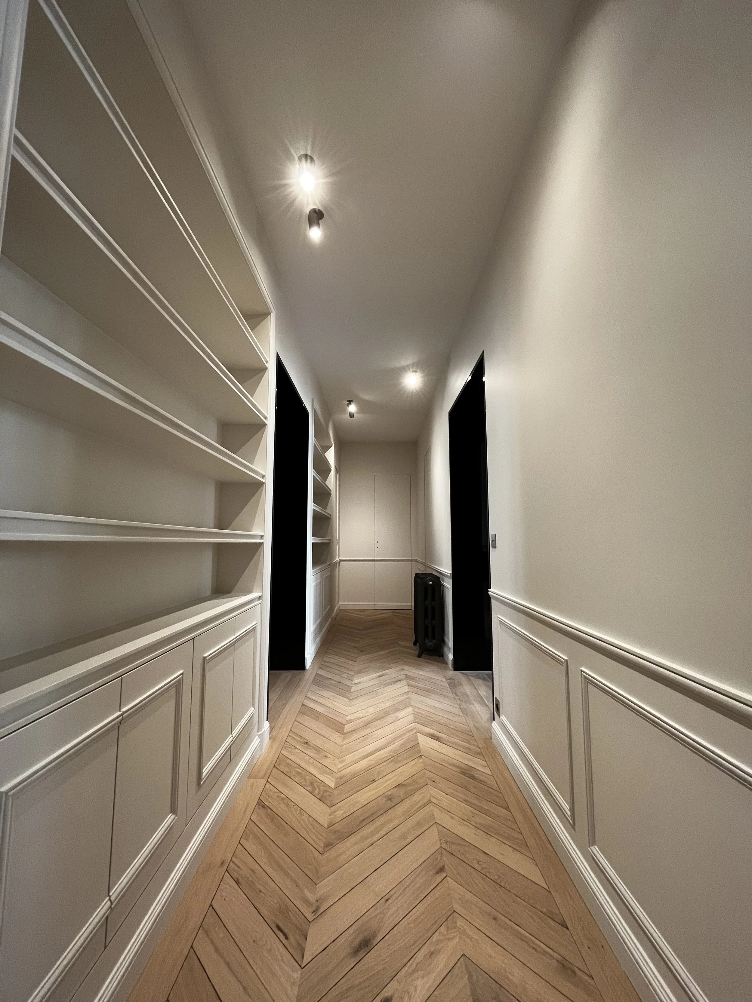 Un couloir intérieur avec des murs blancs, des étagères sur le côté gauche, un parquet en bois clair en motif chevron, et un plafond avec plusieurs spots lumineux.