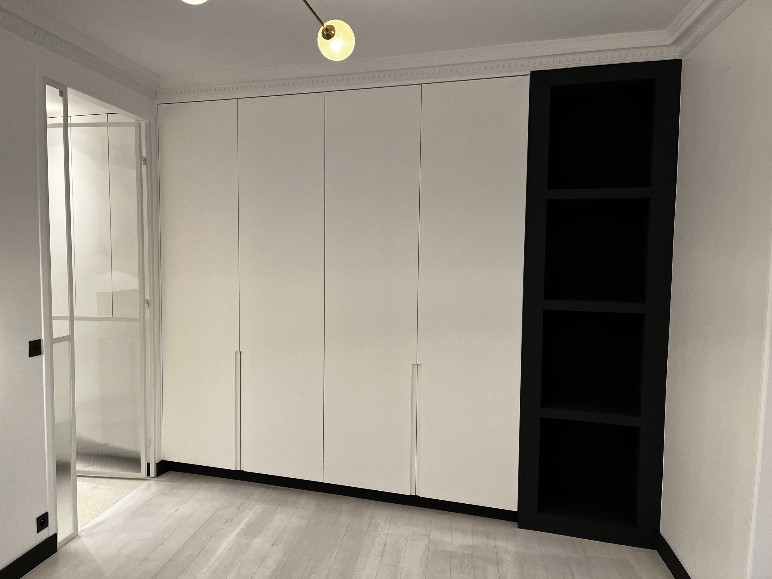 Un grand placard blanc avec des portes à ouverture vers le haut, situé dans une pièce avec des murs blancs, un plafond avec moulures et un sol en bois clair. À gauche, une porte en verre entièrement vitrée s'ouvre sur une autre pièce.