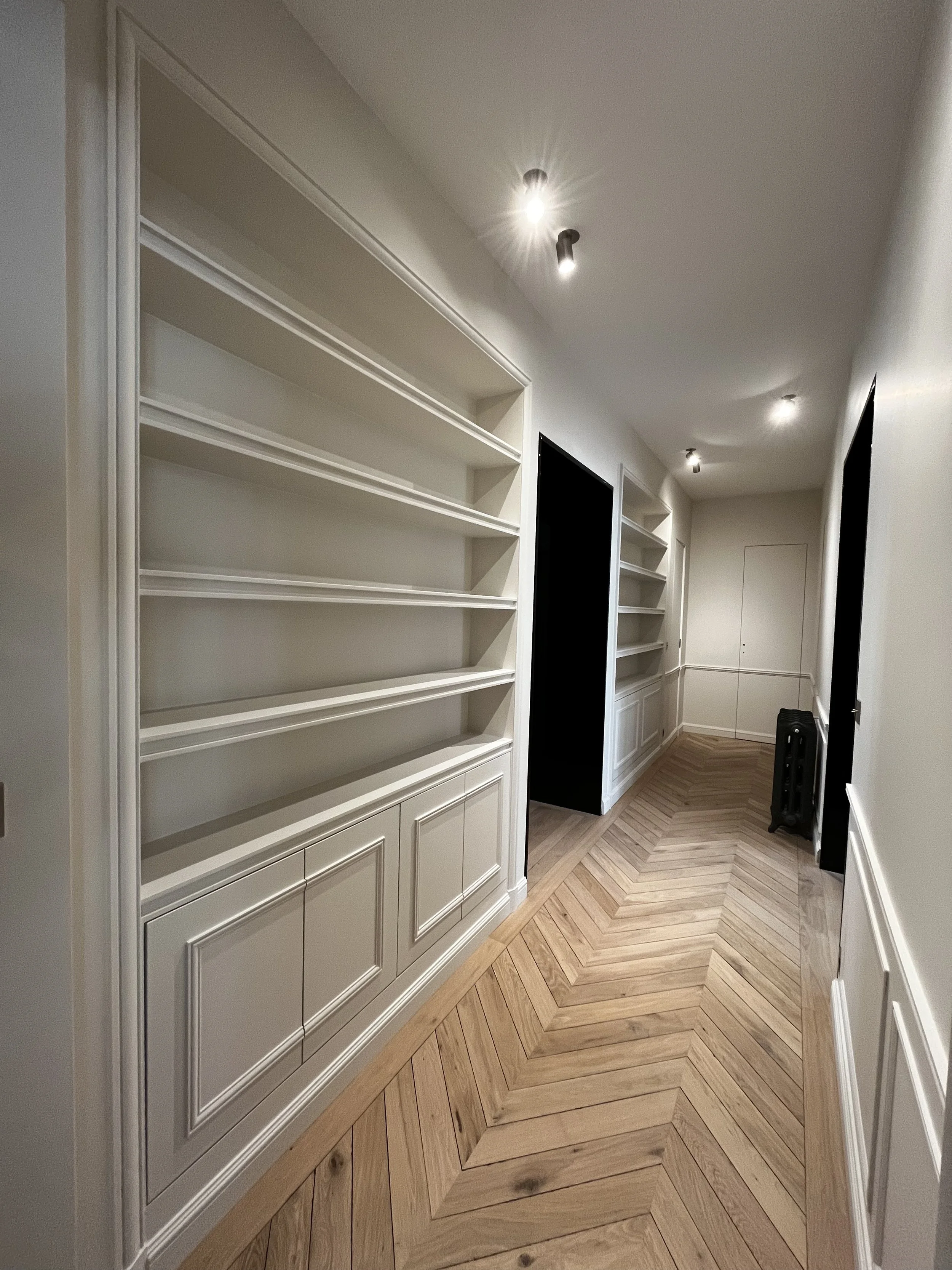 Un couloir intérieur avec des étagères blanches à gauche, un parquet en chevrons en bois clair, et plusieurs spots lumineux au plafond.