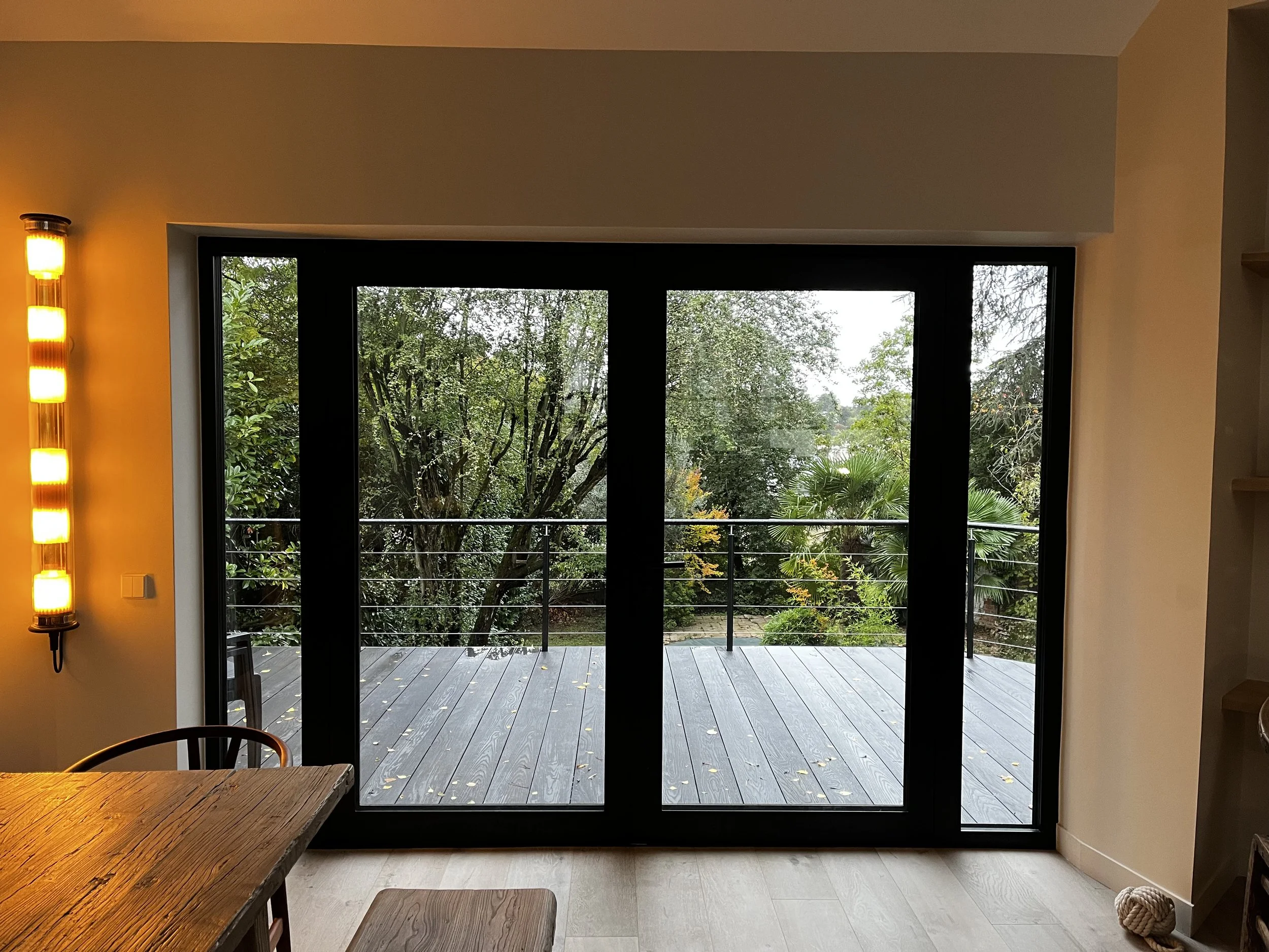 Vue d'une pièce intérieure avec une porte-fenêtre donnant sur un patio en bois. À gauche, un mur avec un éclairage mural à bandes jaunes. Au premier plan, une table en bois avec des chaises.
