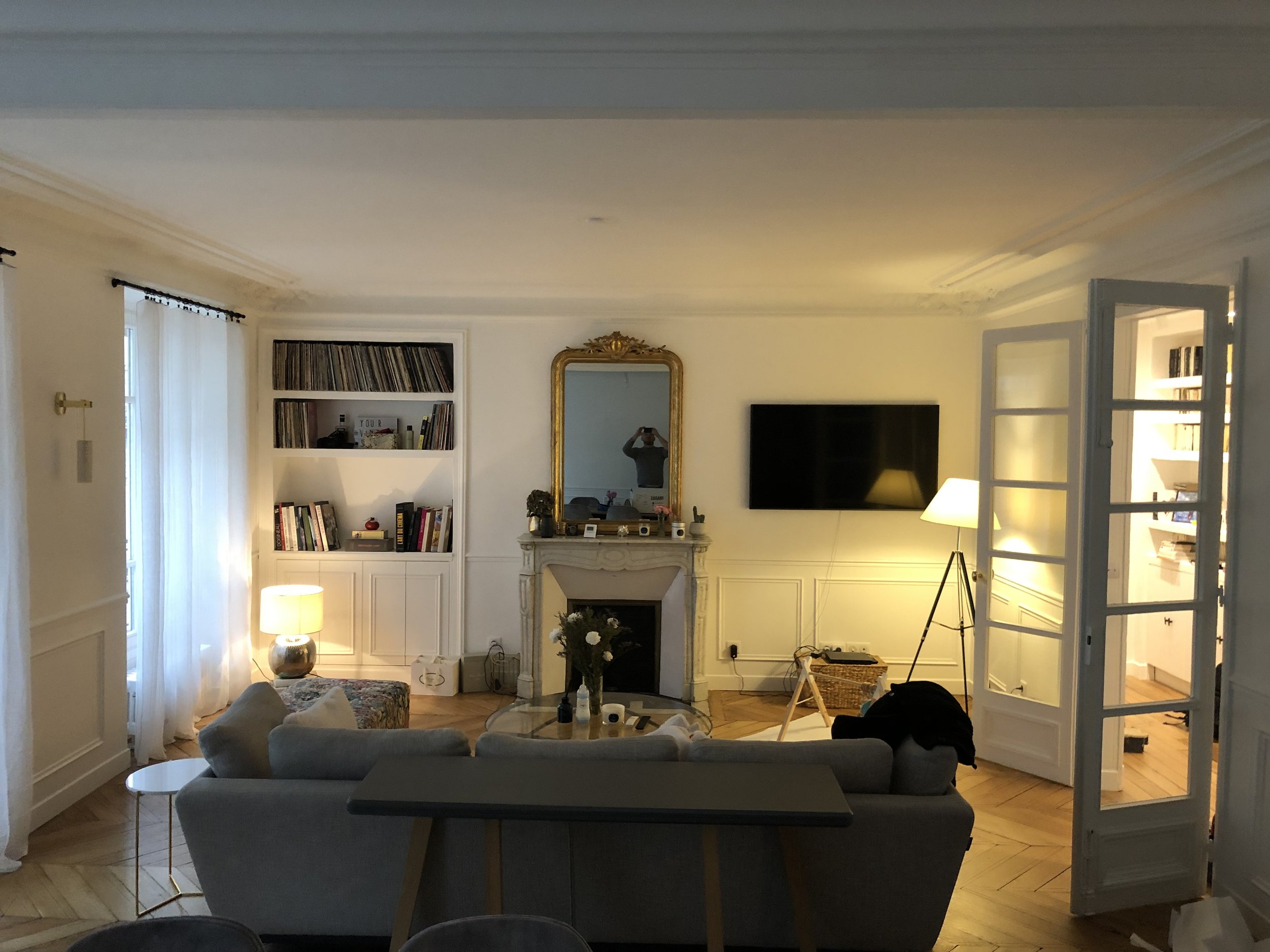 Salon intérieur avec canapé gris, cheminée, grand miroir doré, télévision, lampes et livres.