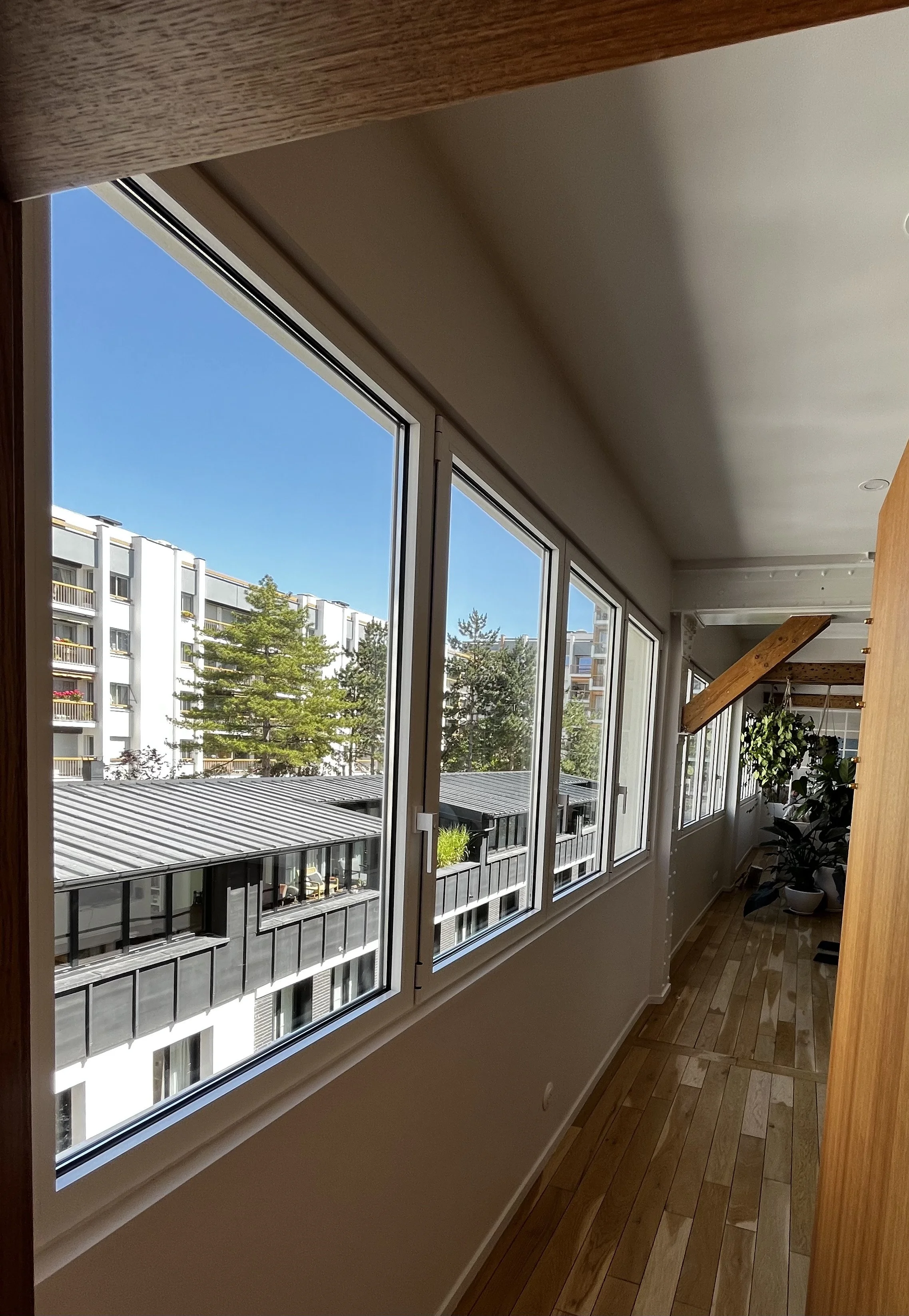 Une vue d'une pièce intérieure avec de grandes fenêtres donnant sur un bâtiment d'appartements entouré d'arbres. La pièce a un sol en bois clair, avec des plantes en pot et un lit en bois visible.