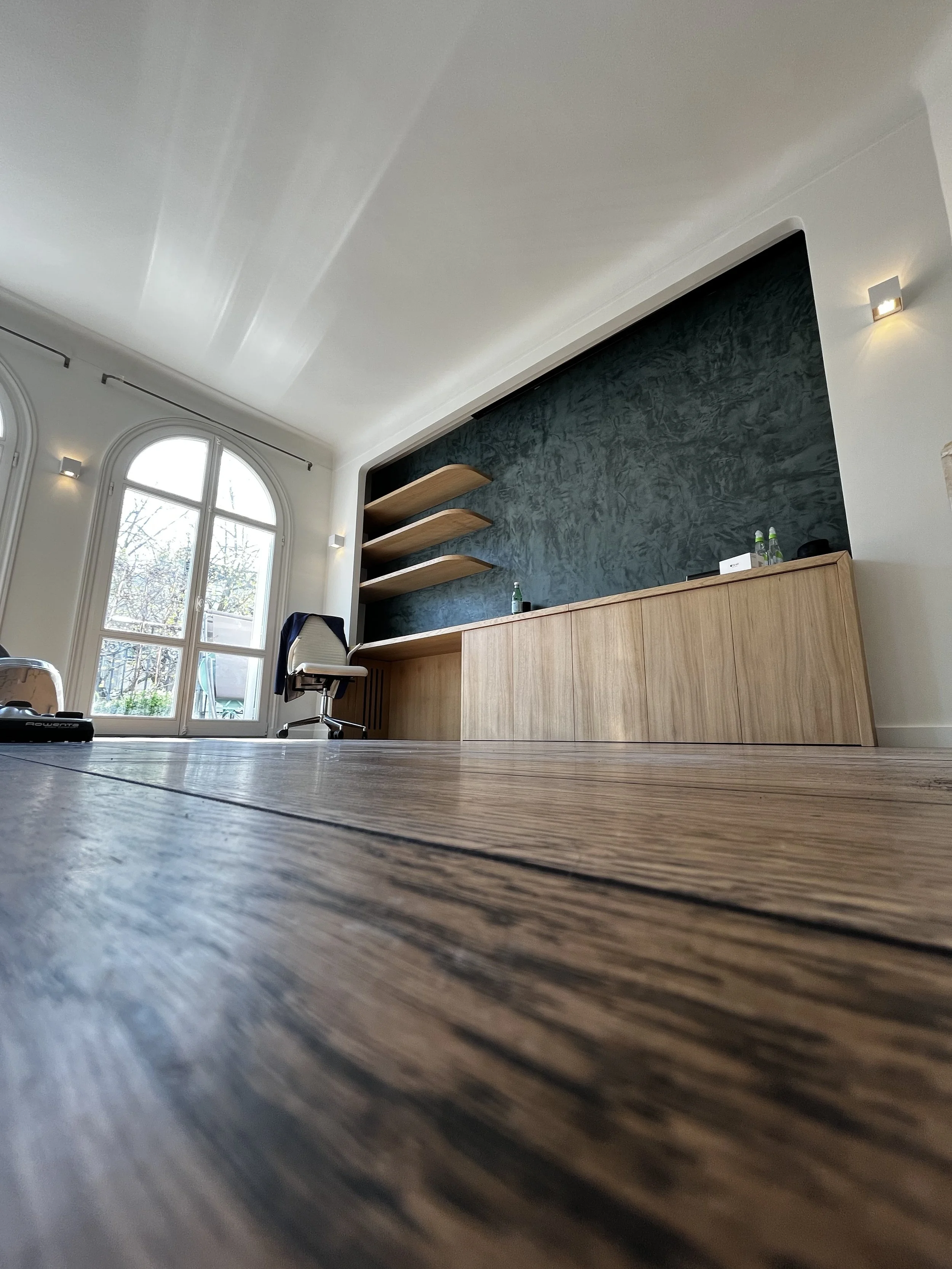 Une pièce moderne avec un sol en bois clair, une grande fenêtre avec volets et une bibliothèque en bois avec des étagères flottantes noires et un meuble de rangement en bois. Il y a quelques bouteilles d'eau sur le meuble.