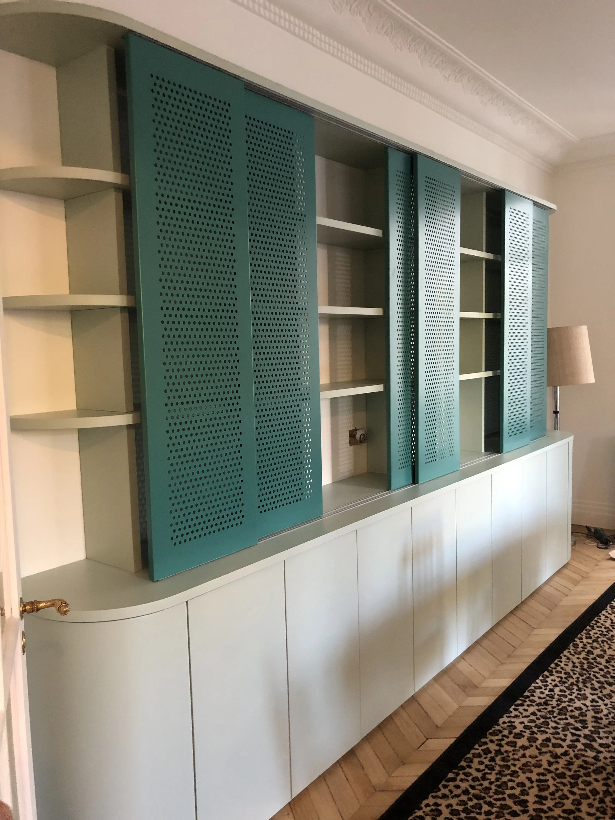 Une grande bibliothèque ou unité de rangement avec des portes perforées turquoise et des étagères vides, située dans une pièce avec parquet en bois clair, tapis à motif léopard, et une lampe à pied à droite.
