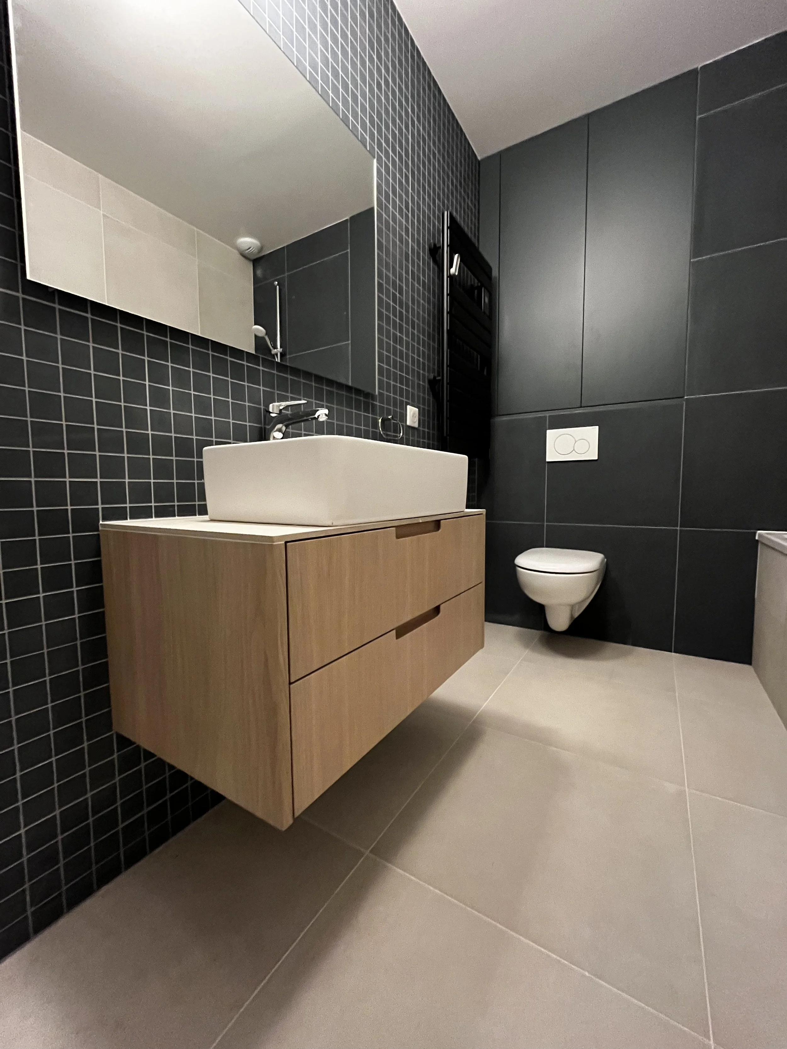 Un lavabo blanc posé sur un meuble en bois clair, dans une salle de bain contemporaine avec murs en carrelage noir et sol en carrelage beige.