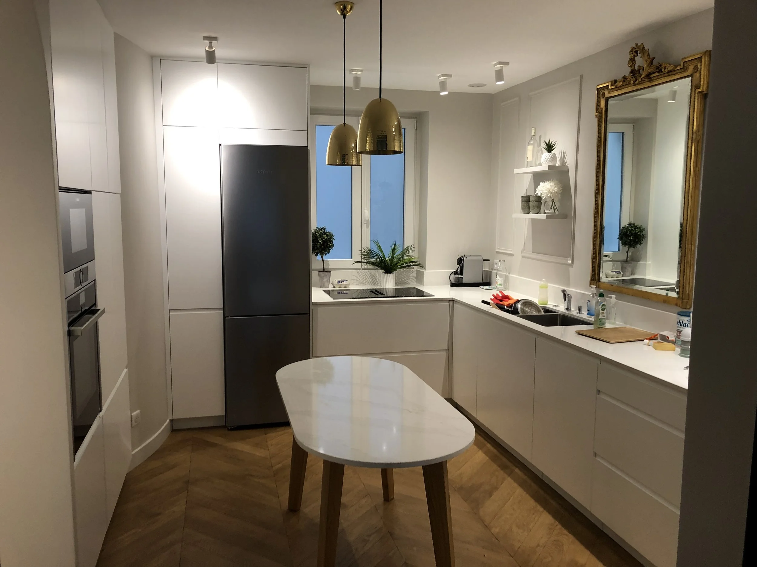 Cuisine moderne avec plan de travail blanc, réfrigérateur en acier inoxydable, grand miroir avec cadre doré, étagères décoratives avec plantes et objets en céramique, lampe suspendue dorée, évier avec produits de nettoyage, et fenêtres laissant entre