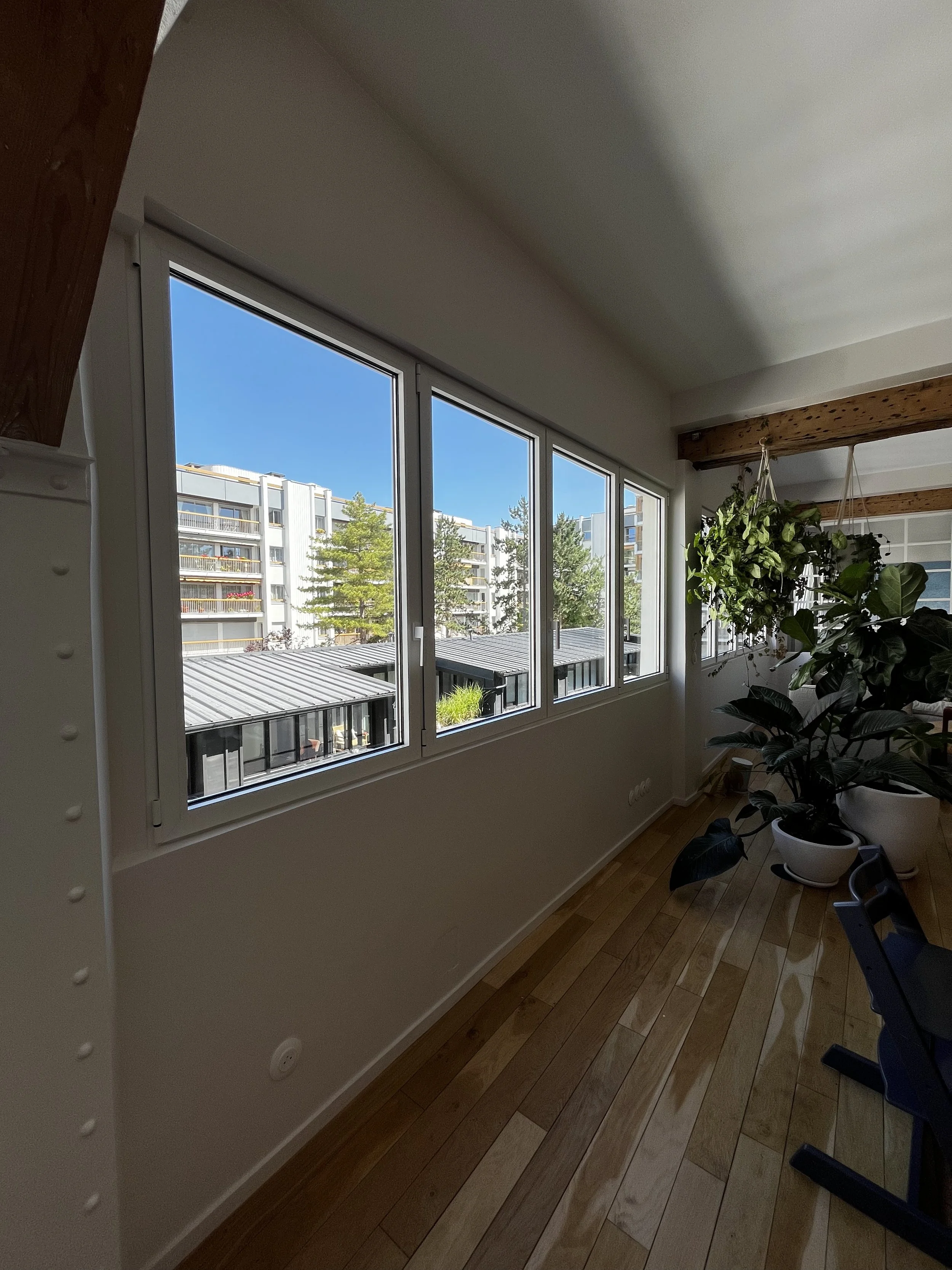 Vue intérieure d'une pièce avec un long mur de fenêtres, des plantes en pot, un plafond à poutres en bois, et un sol en parquet, avec un bâtiment d'immeuble visible à l'extérieur.