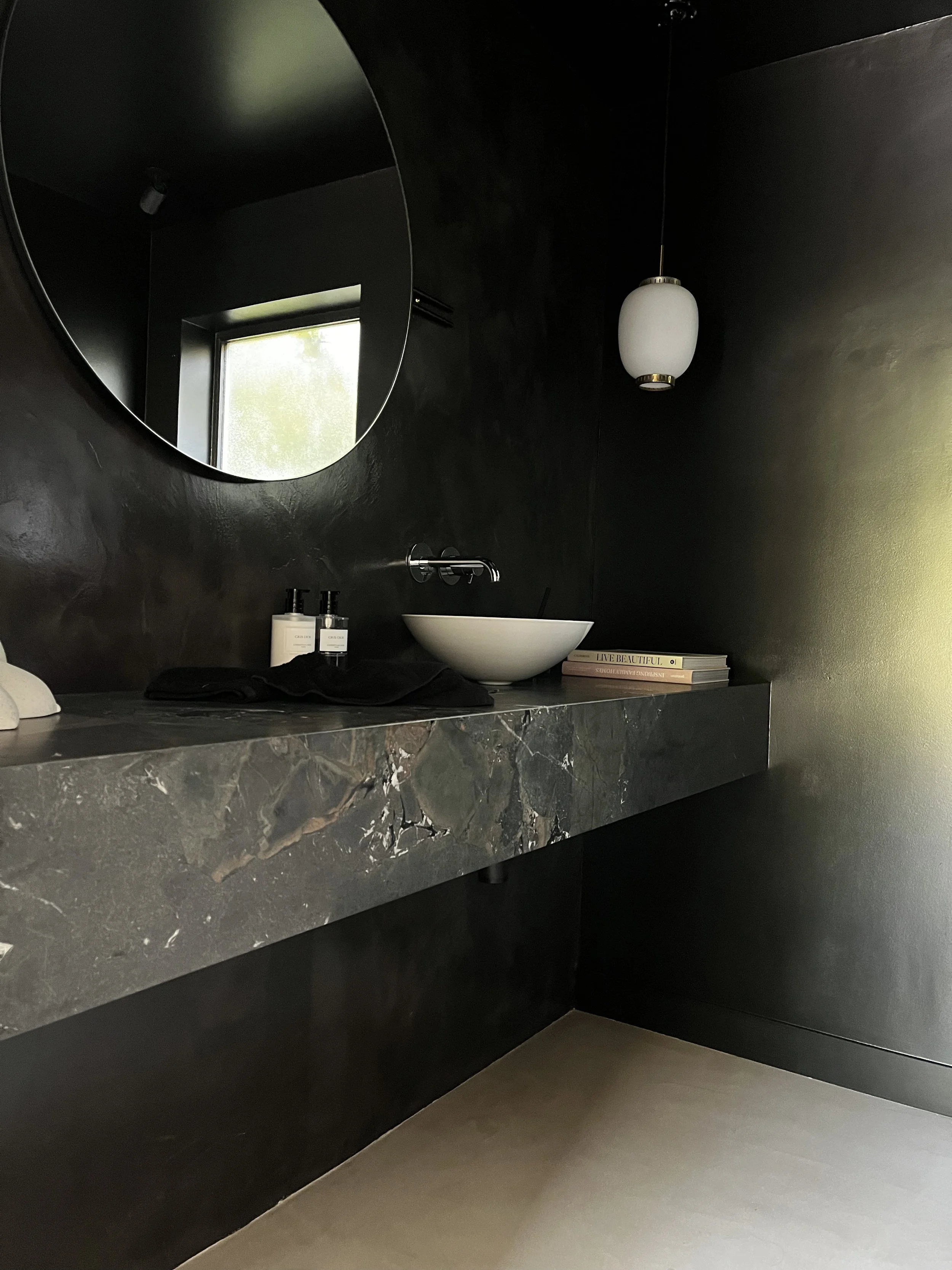 Un lavabo blanc suspendu au mur noir avec un miroir rond au-dessus, une étagère avec des livres et des produits de soins, et une lampe suspendue en forme d'ovale blanc.