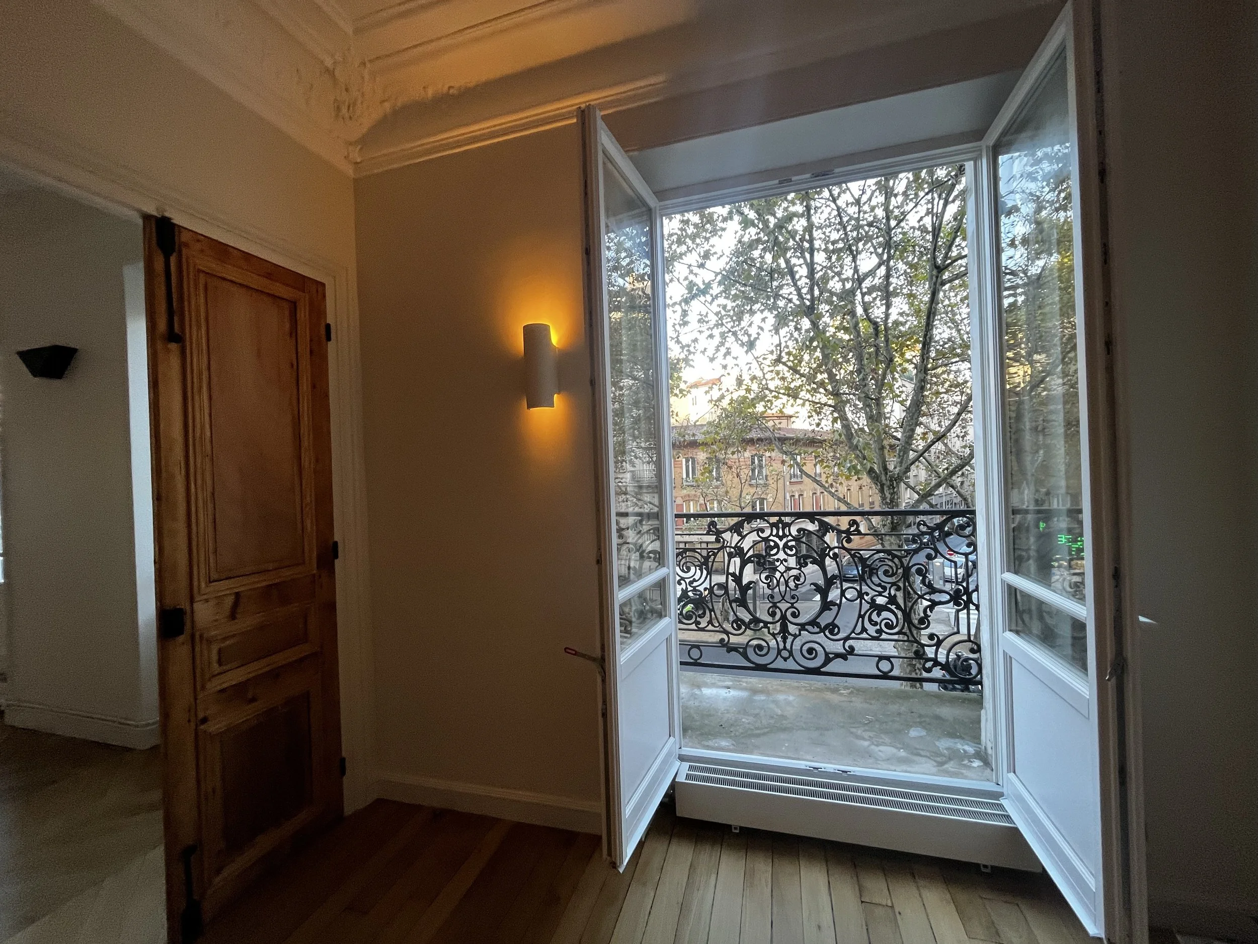 Une porte-fenêtre ouverte donnant sur un balcon avec un motif en fer forgé, laissant entrer la lumière naturelle d'un arbre visible à l'extérieur, dans une pièce avec un mur clair, un lampadaire mural et une porte en bois.
