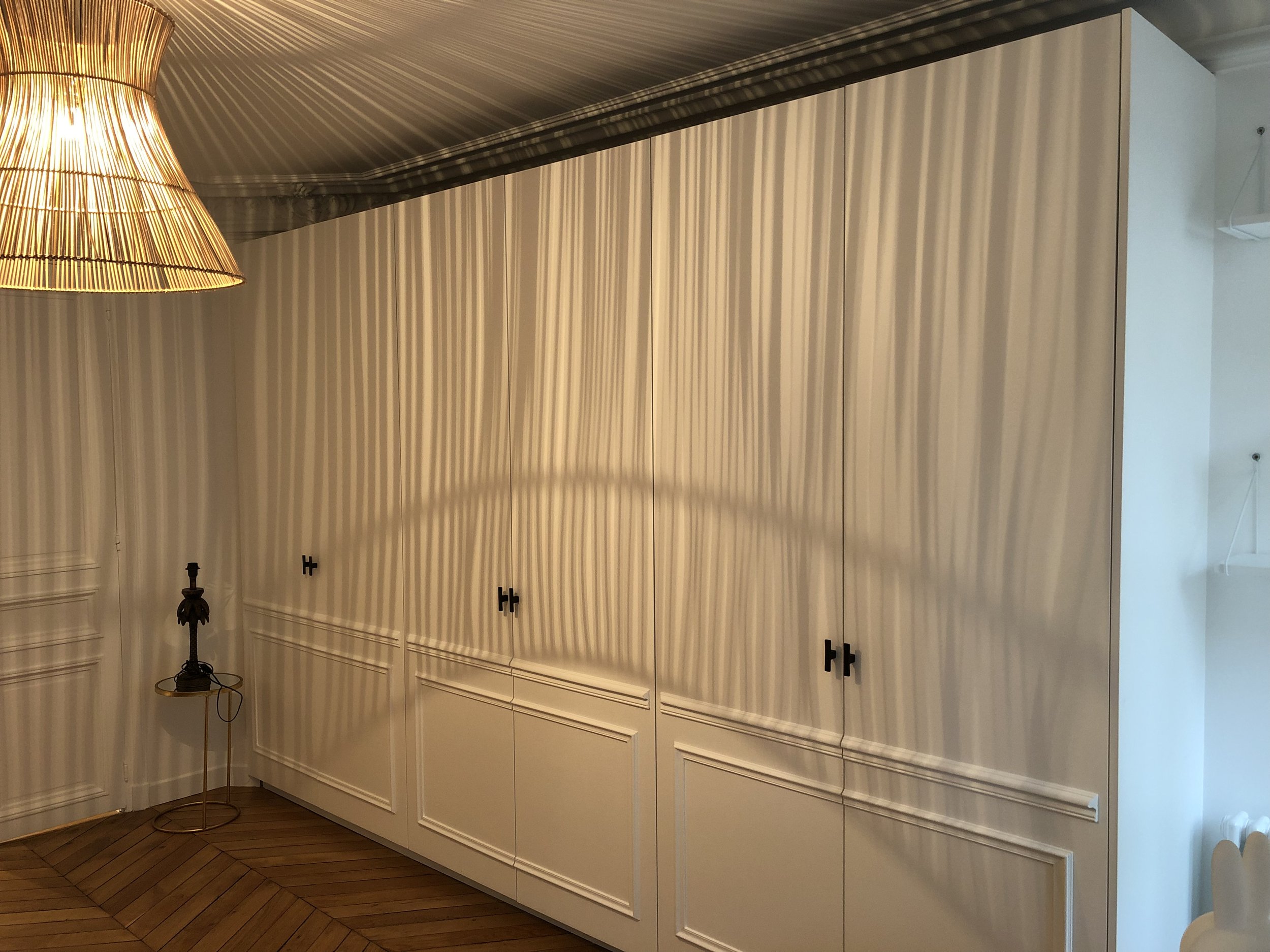 Une grande armoire blanche avec des portes en verre dans une pièce bien éclairée. À gauche, une lampe en bambou émet une lumière chaude, posée sur une petite table en or. Le sol est en parquet en bois foncé.