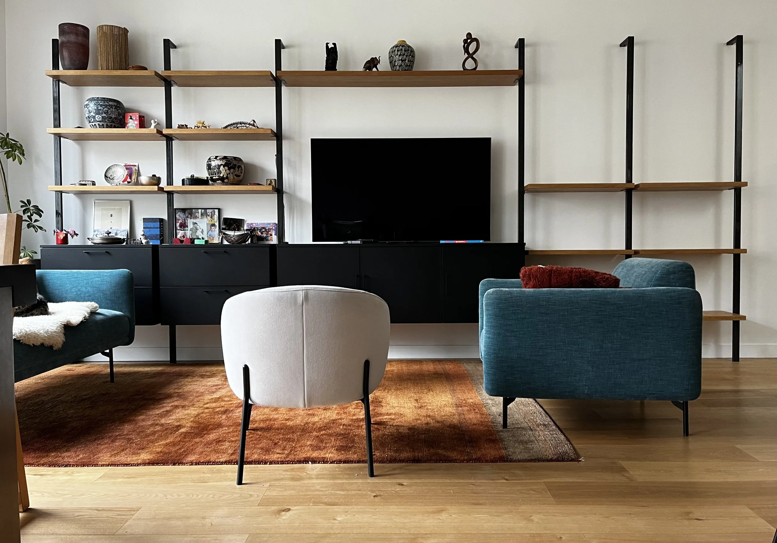 Salon avec étagères en bois et métal contenant des objets décoratifs, télévision au centre, assises en tissu bleu et blanc, tapis en dégradé de rouge et orange, et parquet en bois clair.