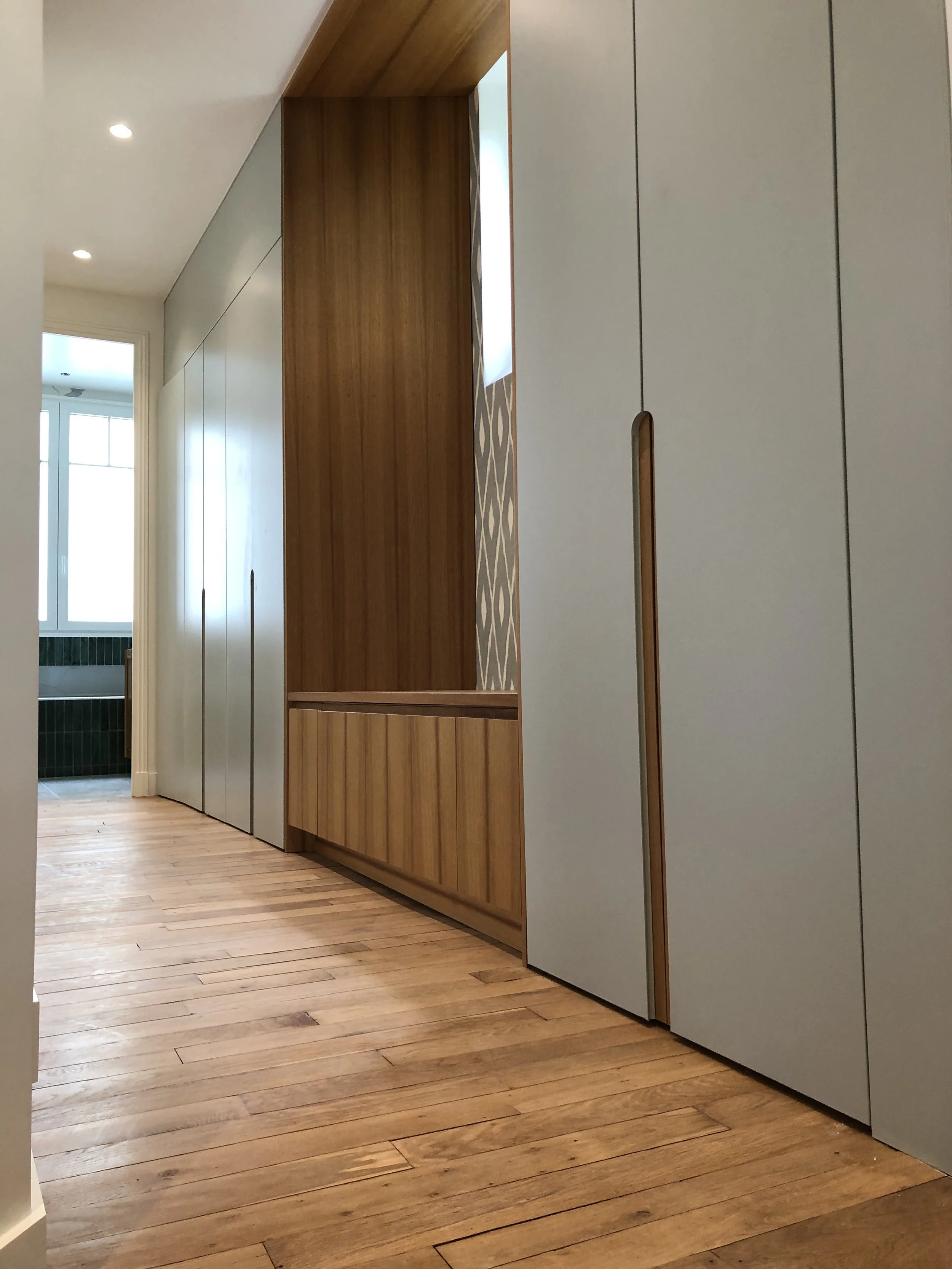 Un couloir avec un plancher en bois clair, des murs gris et un grand placard avec des portes grises, une pièce lumineuse en arrière-plan