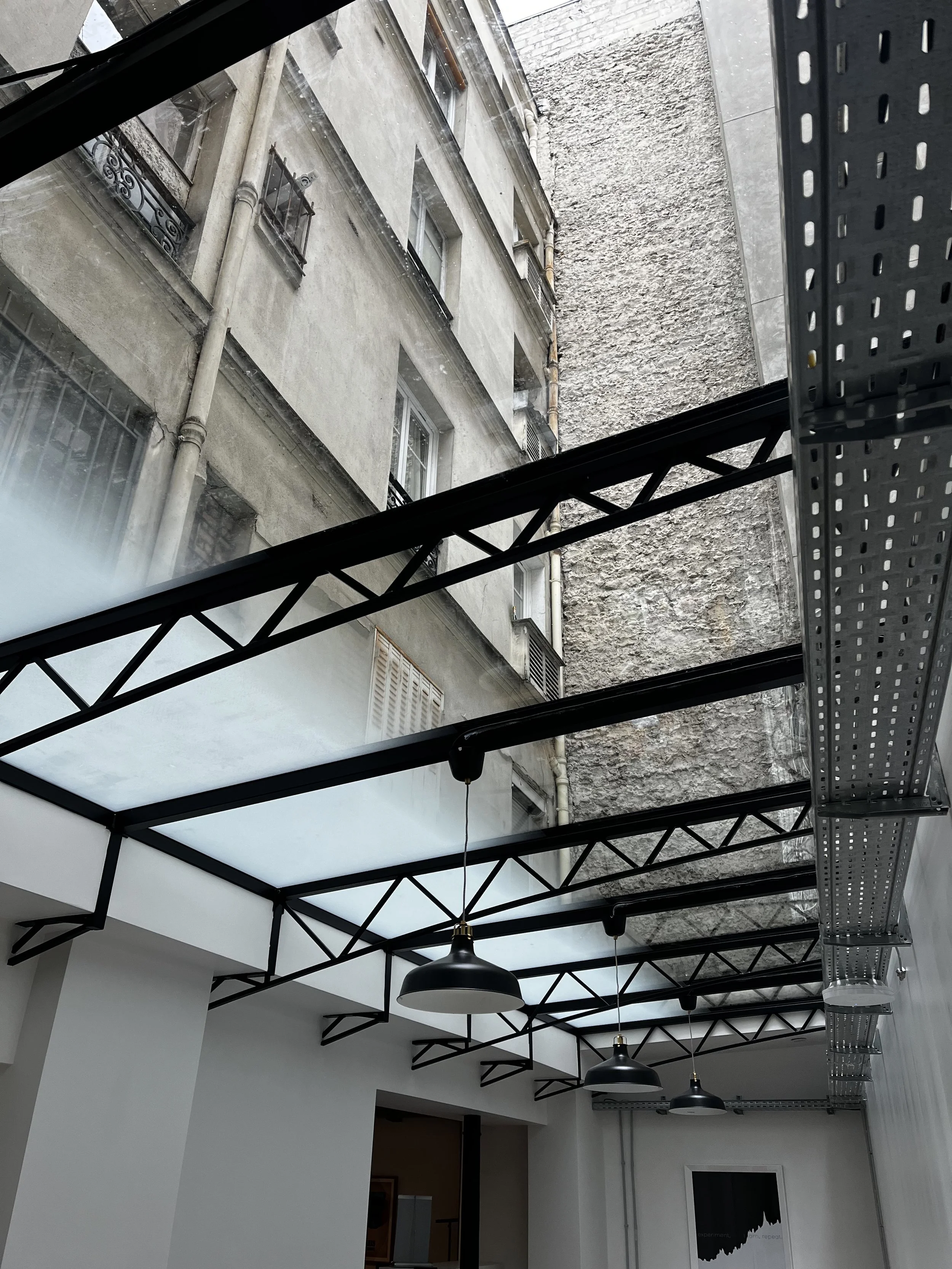 Intérieur d'un espace moderne avec plafond en verre, lampes suspendues, murs blancs et structure métallique noire visible.