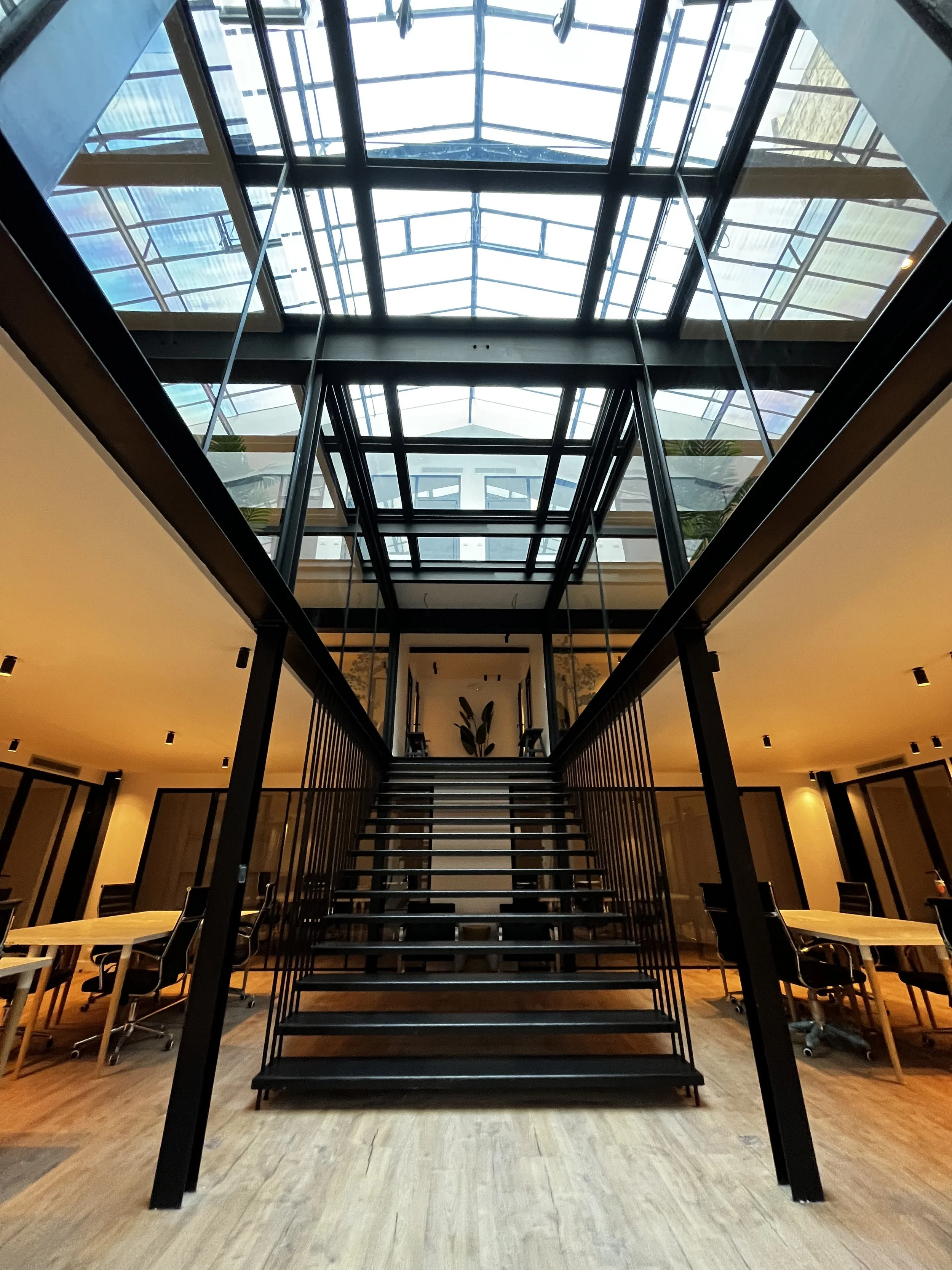 Escalier en métal noir au centre dans une salle avec un plafond en verre à plusieurs étages. Mobilier en bois et chaises autour, ambiance moderne.