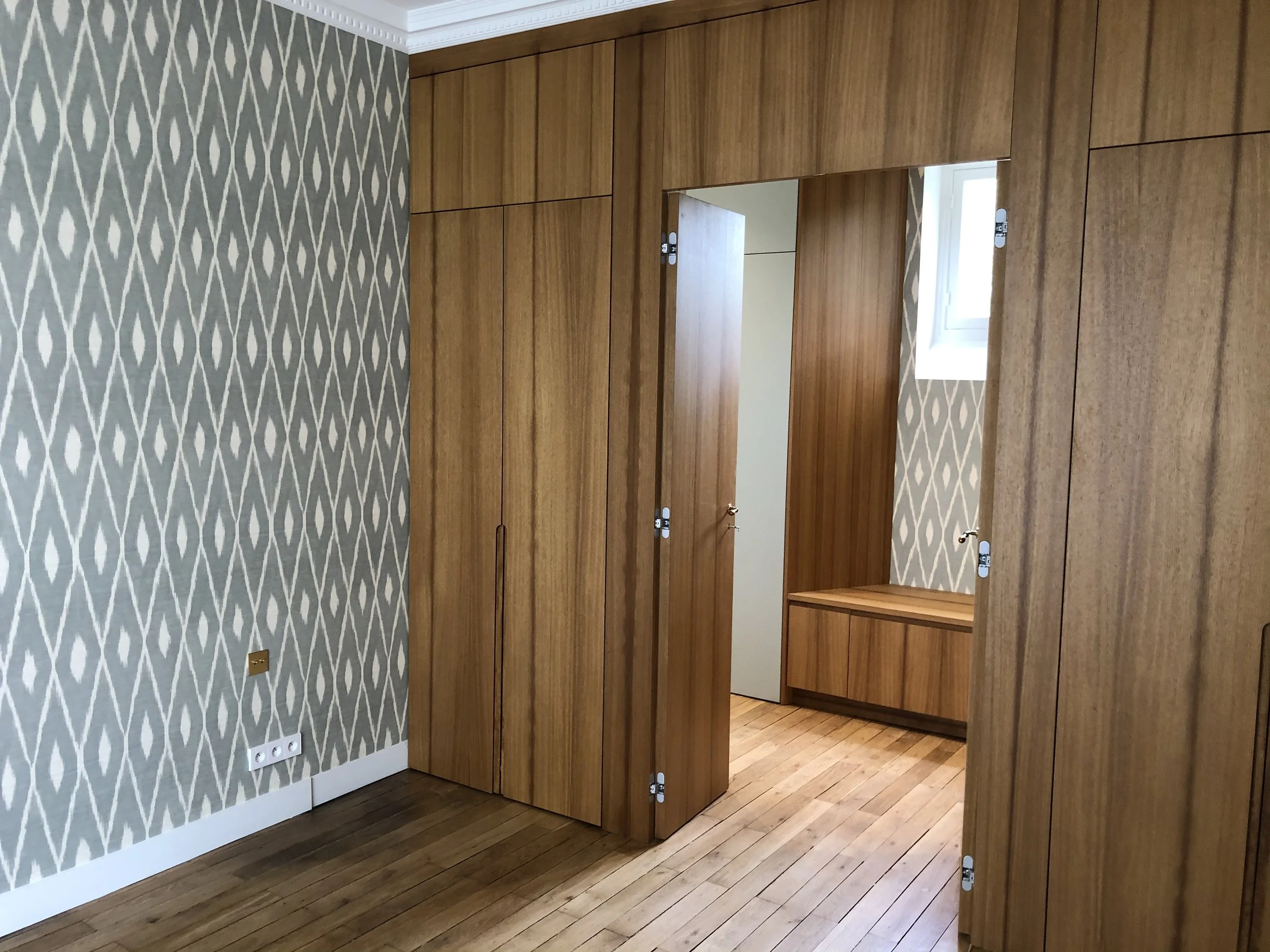 Un mur avec un papier peint à motif géométrique gris et blanc, un grand placard en bois avec portes et miroirs, et un sol en bois clair, dans une pièce intérieure.