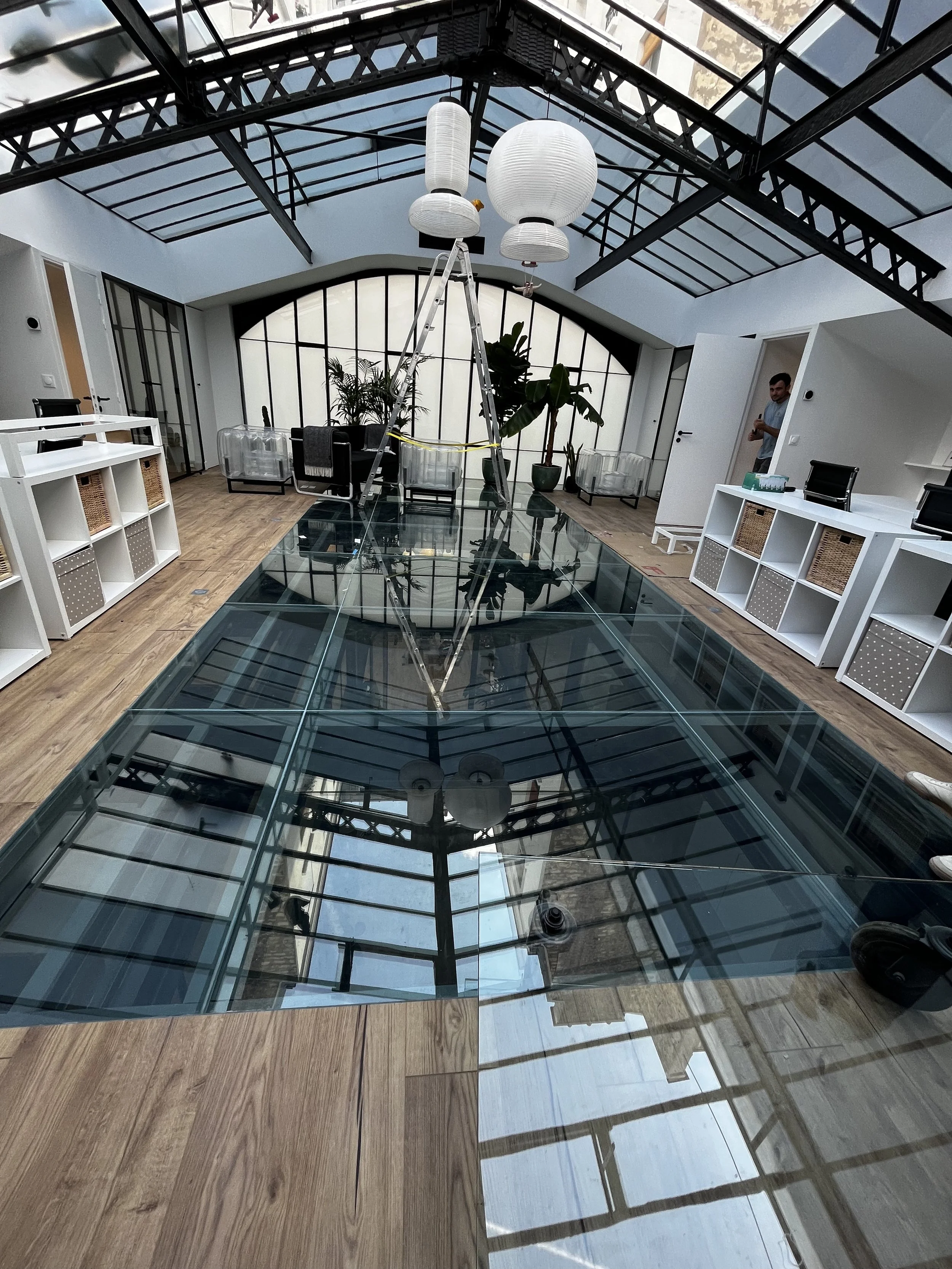 Intérieur d'un espace moderne avec un plafond en verre, une échelle, des lampes en papier blanc suspendues, des plantes vertes, et des meubles blancs.