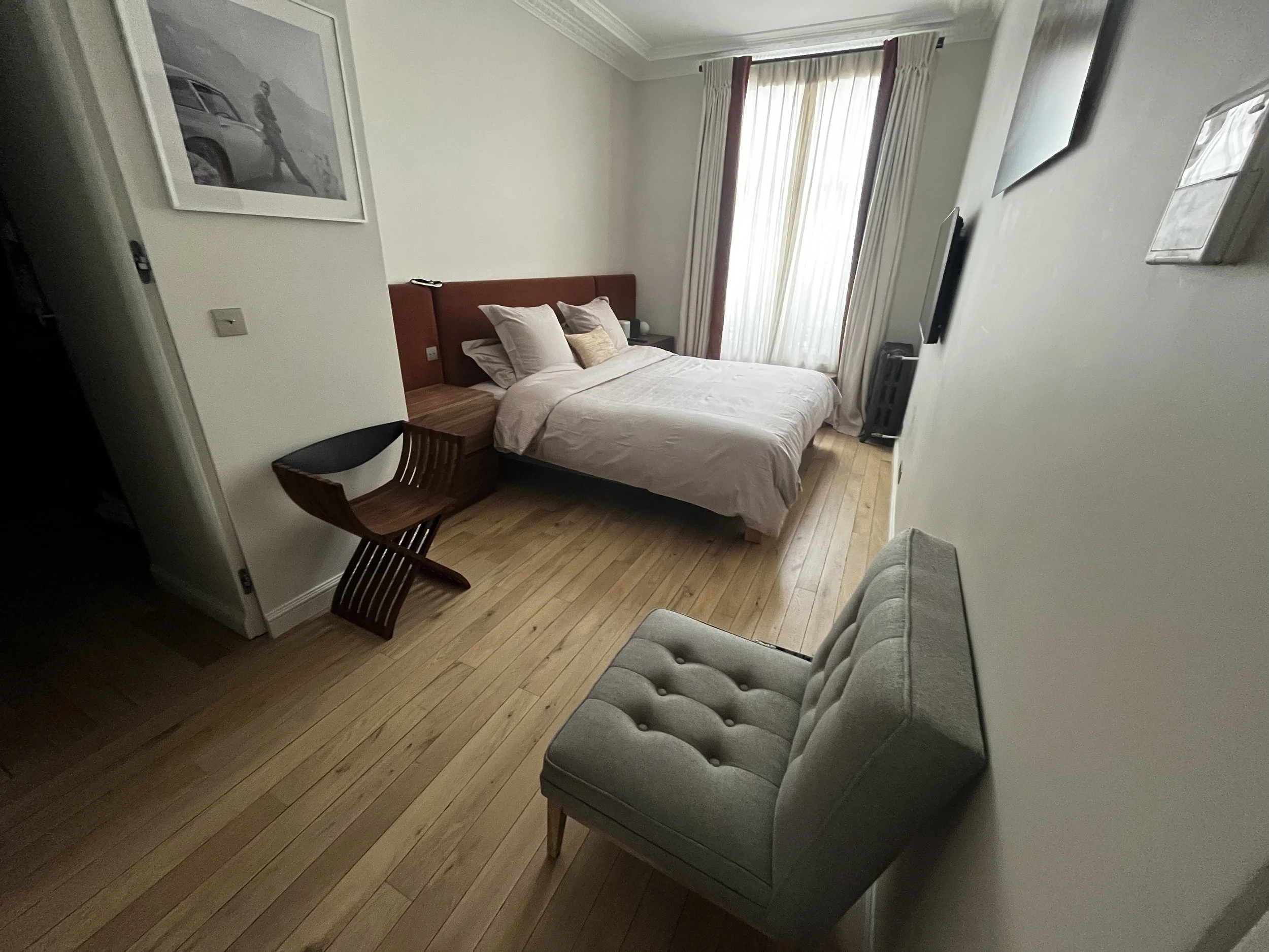 Chambre avec un lit, un fauteuil gris, un tableau au mur, une télévision et de grandes fenêtres avec rideaux.