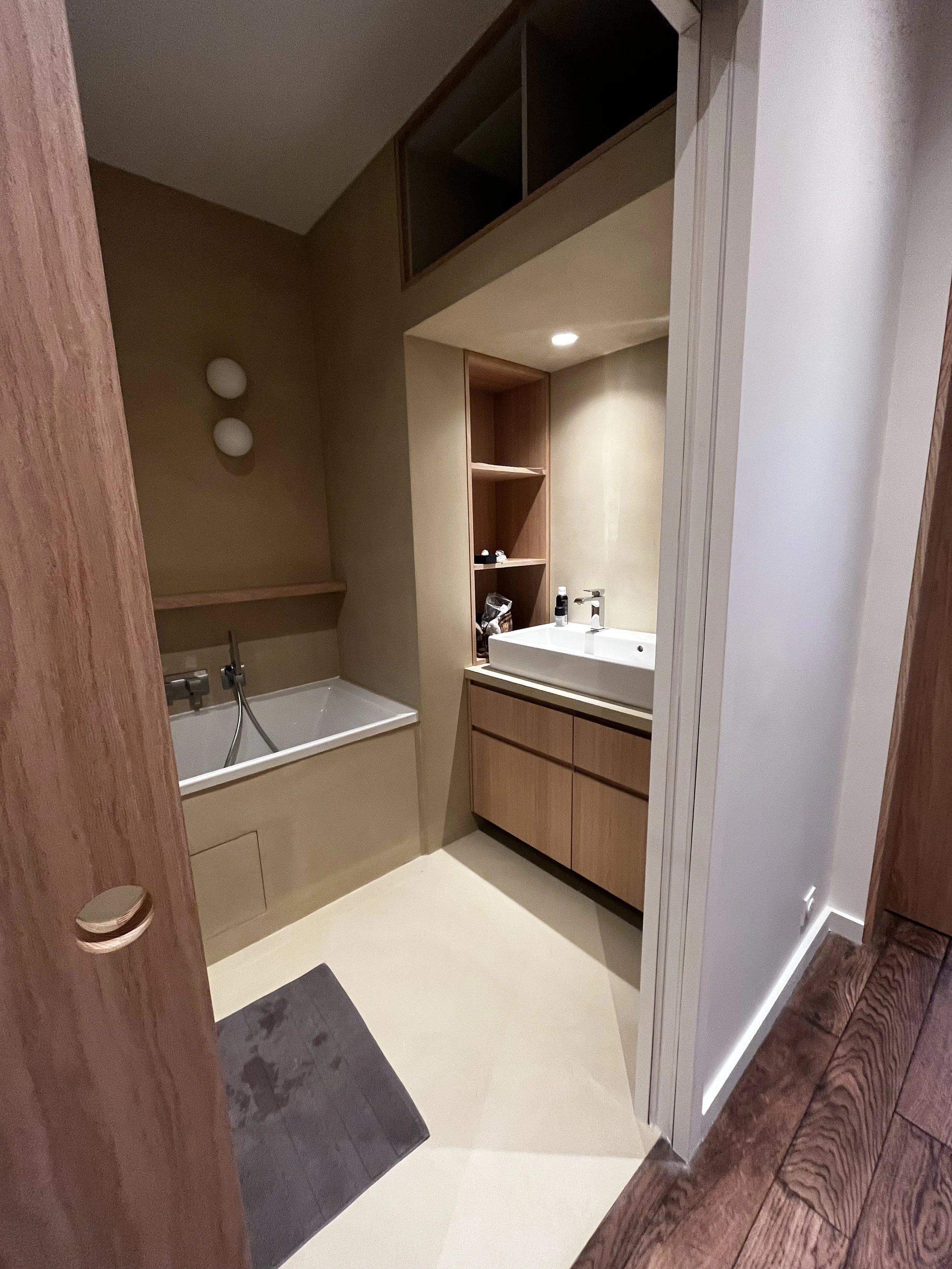 Une salle de bain moderne avec un lavabo, des étagères en bois, un miroir, une baignoire, un miroir intégré, un éclairage encastré au plafond, des murs beige et en bois, et un sol en carreaux gris et beige.