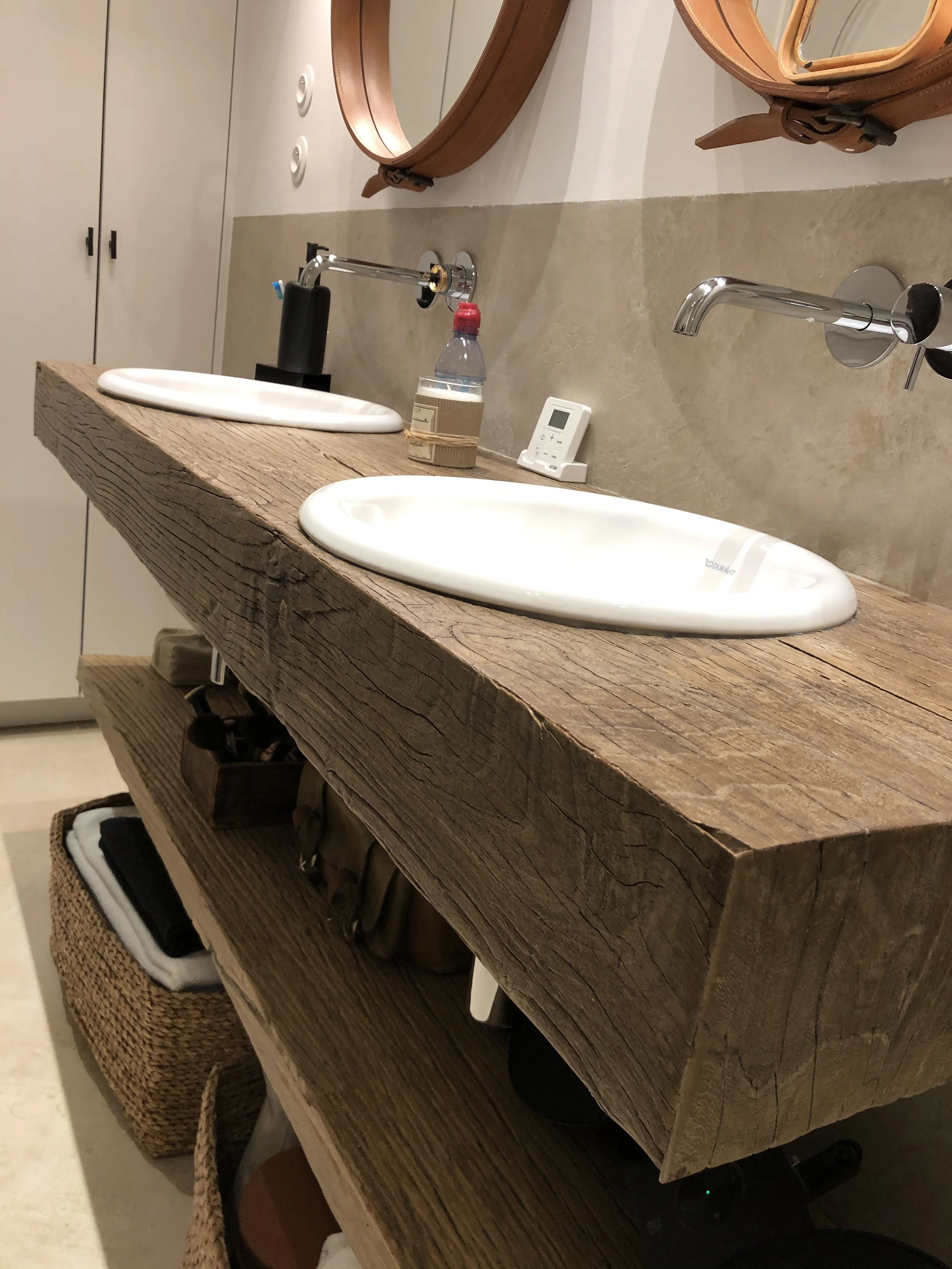 Deux lavabos en céramique blanche sur une surface en bois brut, avec un miroir rond en haut à droite, un distributeur d'eau et un thermostat. Des armoires blanches avec poignées noires en arrière-plan.