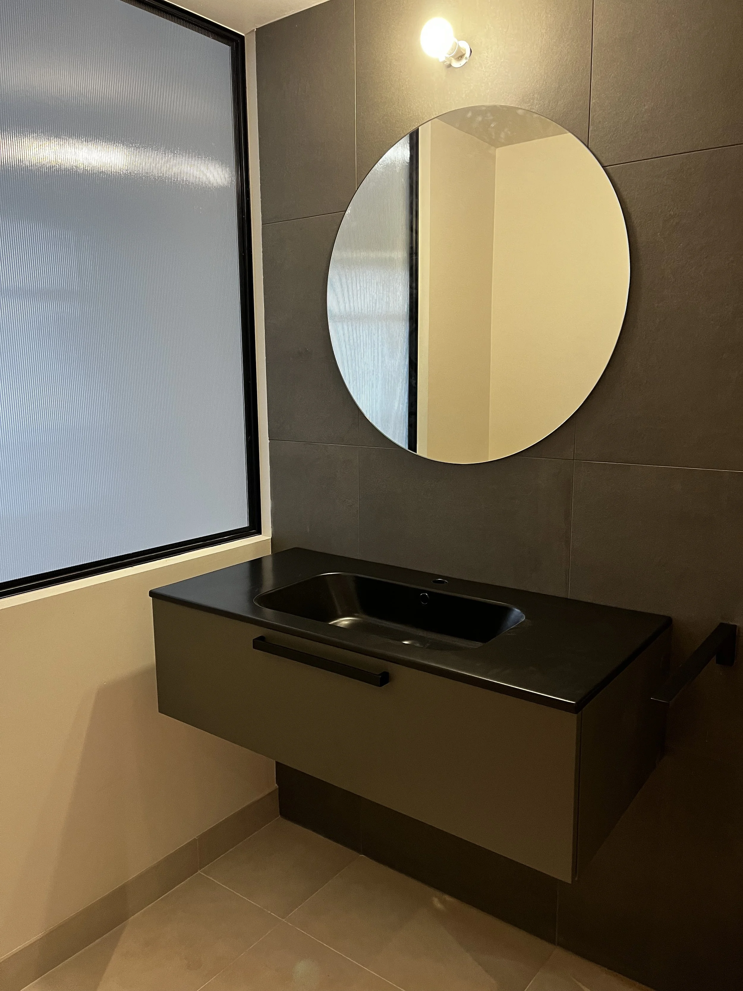 Grand miroir rond fixé au mur au-dessus d'un évier noir dans une salle de bain moderne avec carrelage foncé et une fenêtre avec voilage opaque.